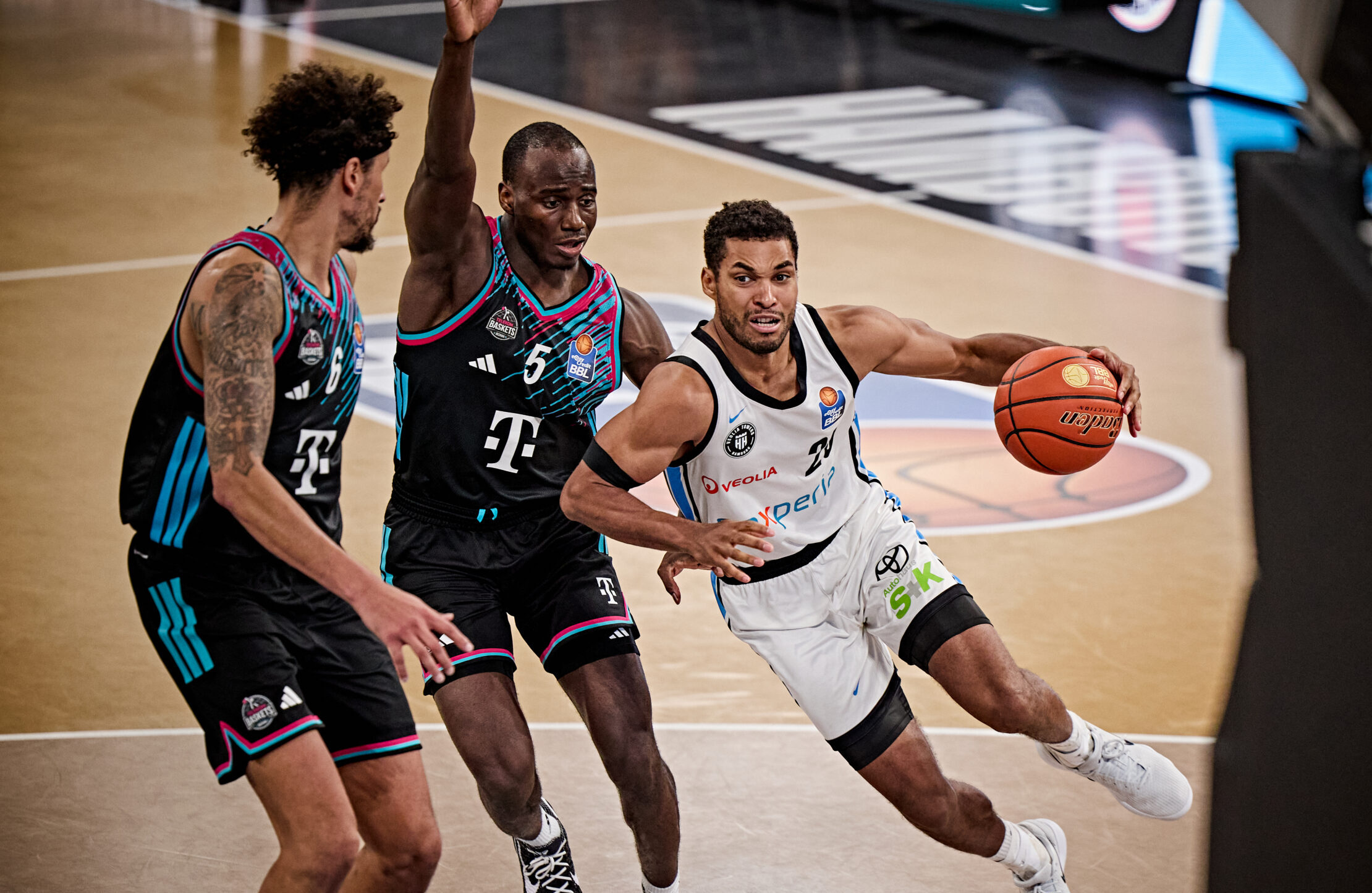 Drei Basketballspieler in Aktion auf dem Spielfeld, einer mit Ball in weißem Trikot dribbelt an zwei Gegenspielern in schwarzen Trikots vorbei