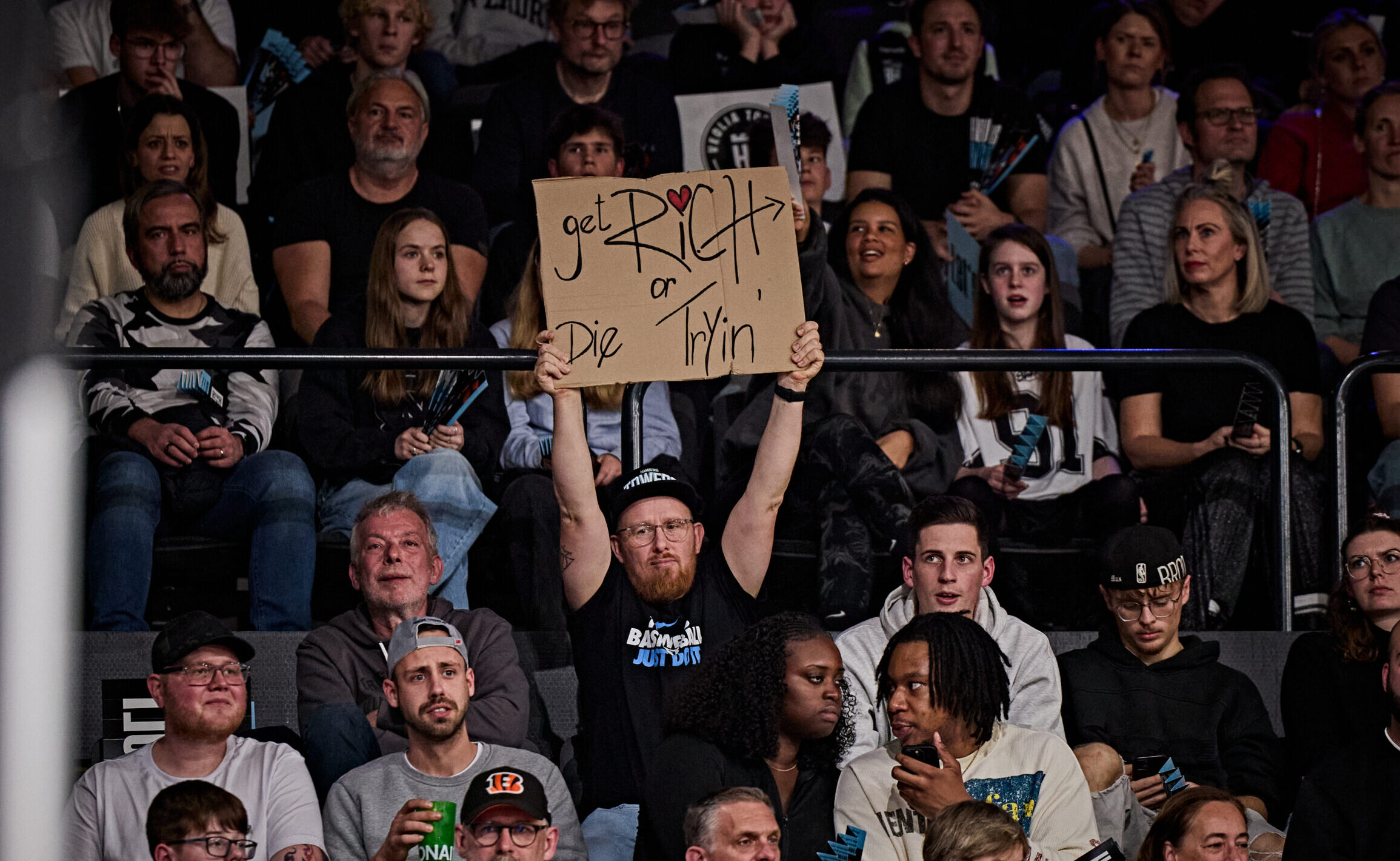 Zuschauer in einer Halle, eine Person hält ein Schild mit der Aufschrift 'get RICH or Die Tryin' hoch