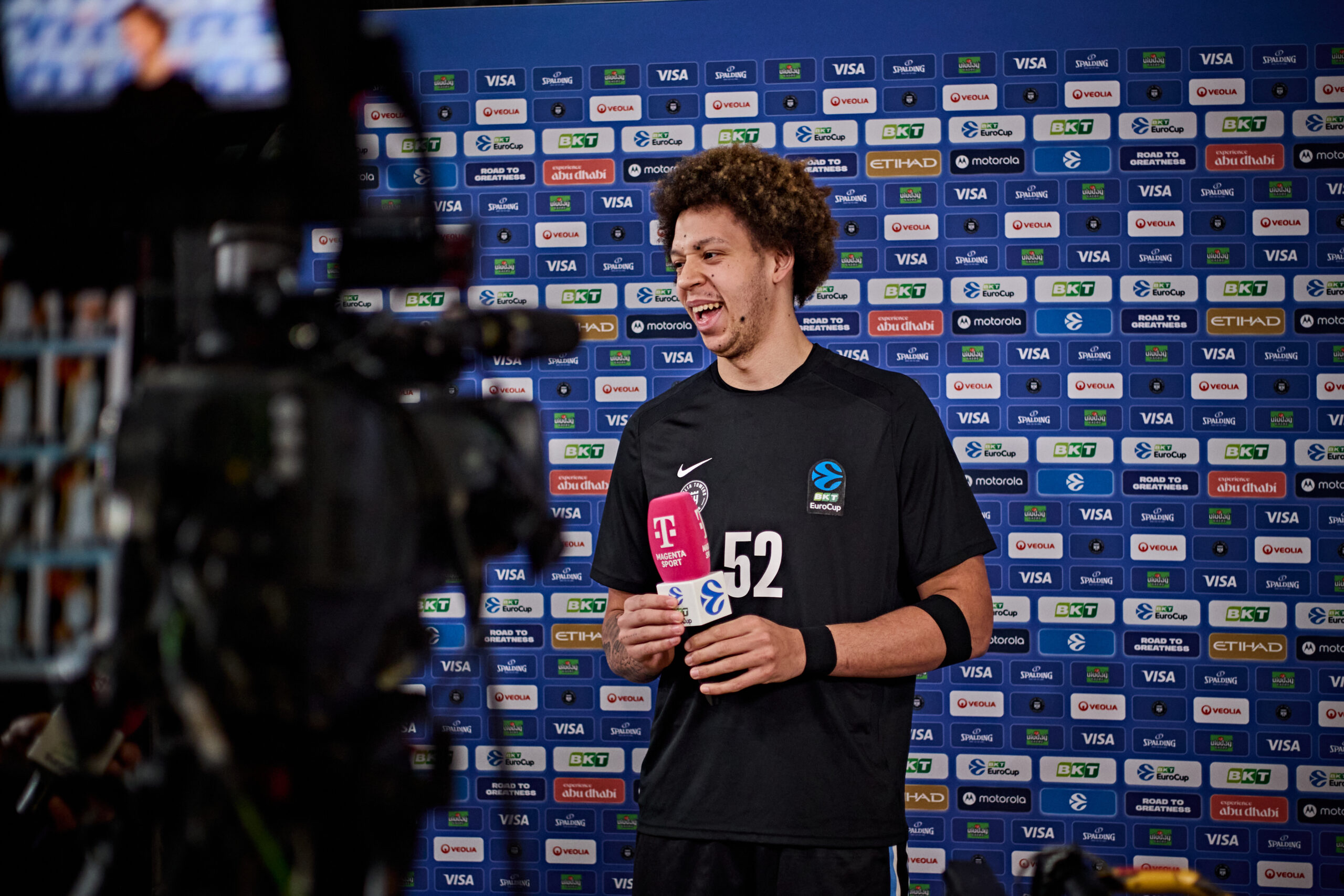 Ein Sportler in schwarzem Trikot mit der Nummer 52 hält ein Mikrofon vor einer Sponsorenwand mit zahlreichen Logos