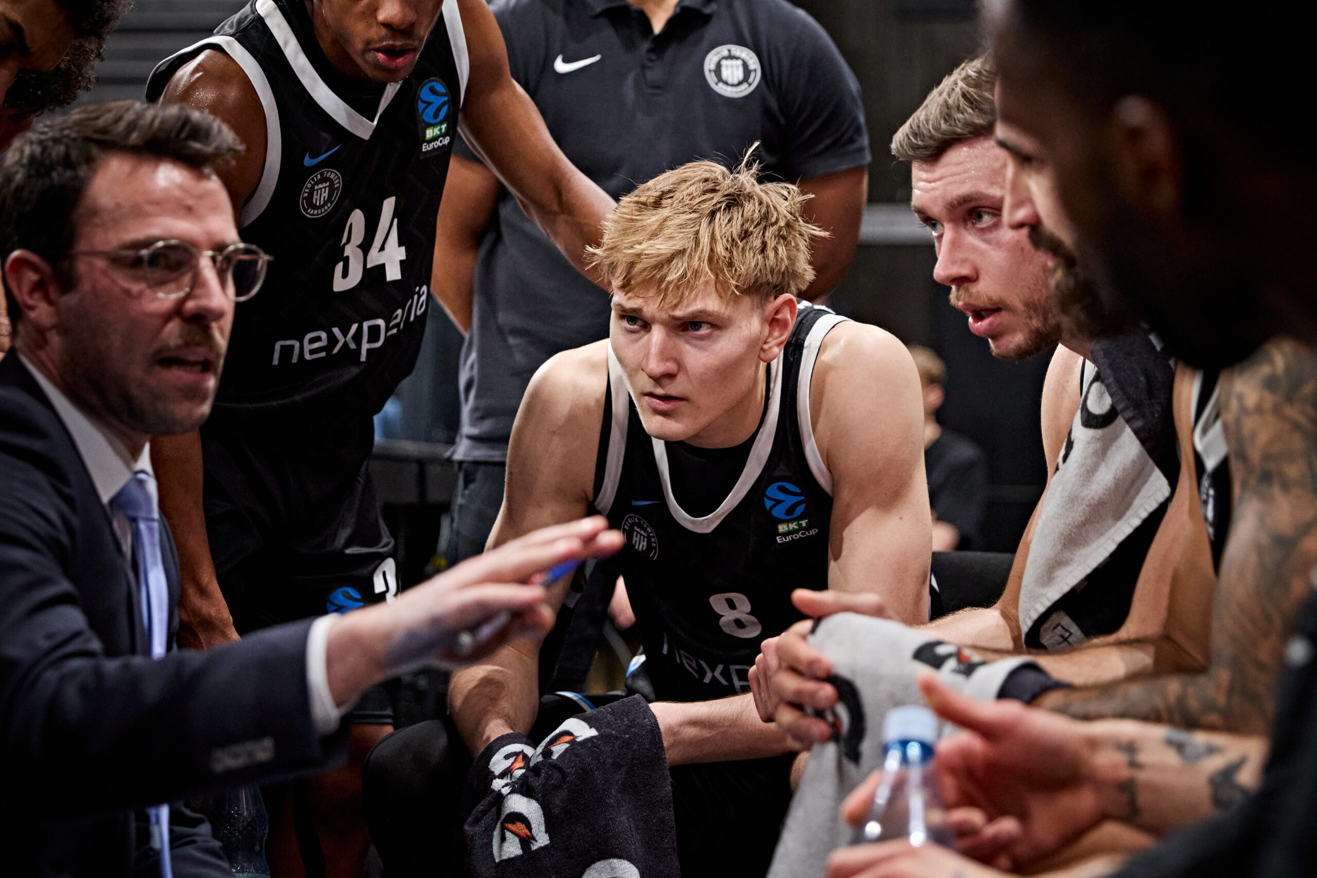 Basketballspieler in schwarzen Trikots mit weißen Details sitzen und stehen in einer Teambesprechung auf der Bank, ein Trainer gestikuliert mit der Hand