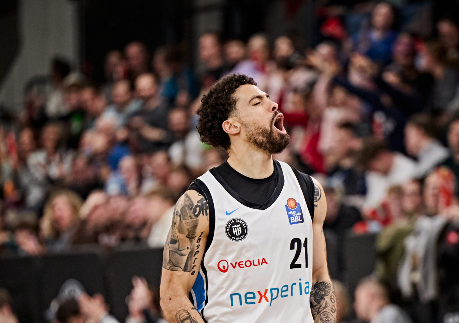 Basketballspieler mit Trikotnummer 21 und Tattoos auf dem linken Arm steht auf dem Spielfeld vor Zuschauern