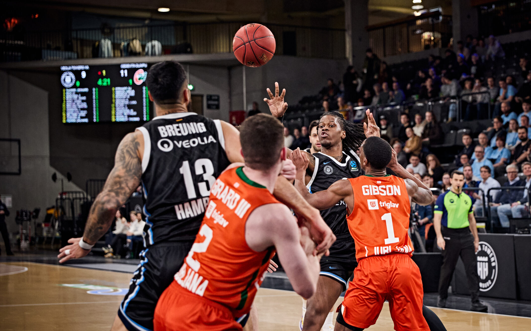 Basketballspieler in orange und schwarz kämpfen um den Ball in einer Halle mit Zuschauern auf den Rängen