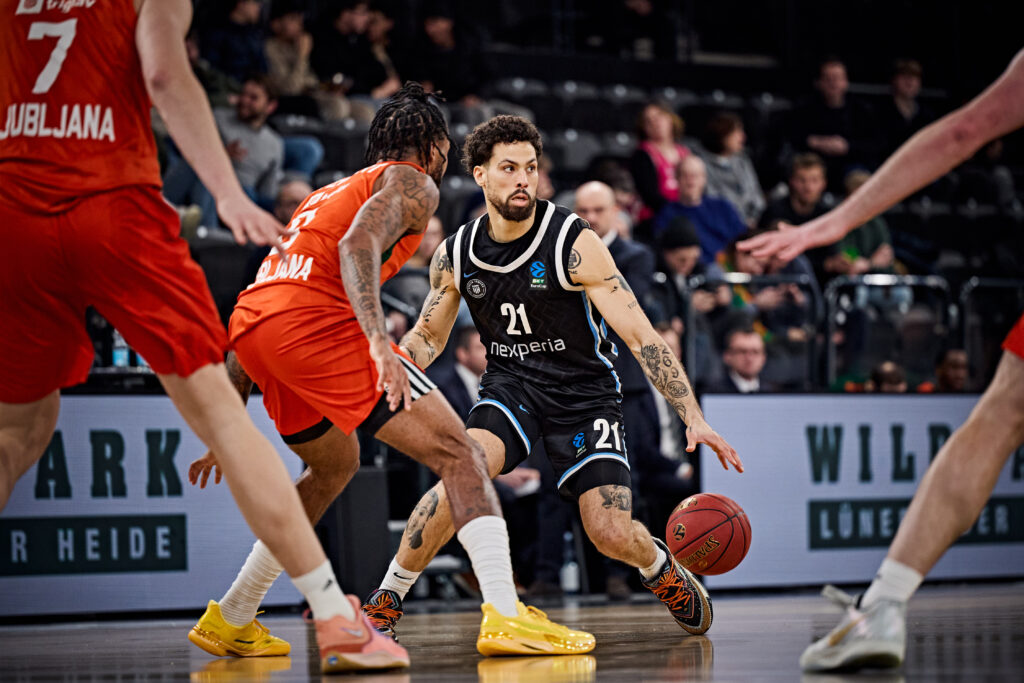 Basketballspieler in schwarzem Trikot mit Ball wird von mehreren Spielern in roten Trikots verteidigt