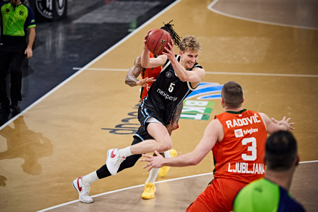 Basketballspieler in schwarzem Trikot mit Ball springt an einem Verteidiger in rotem Trikot vorbei auf einem Hallenboden