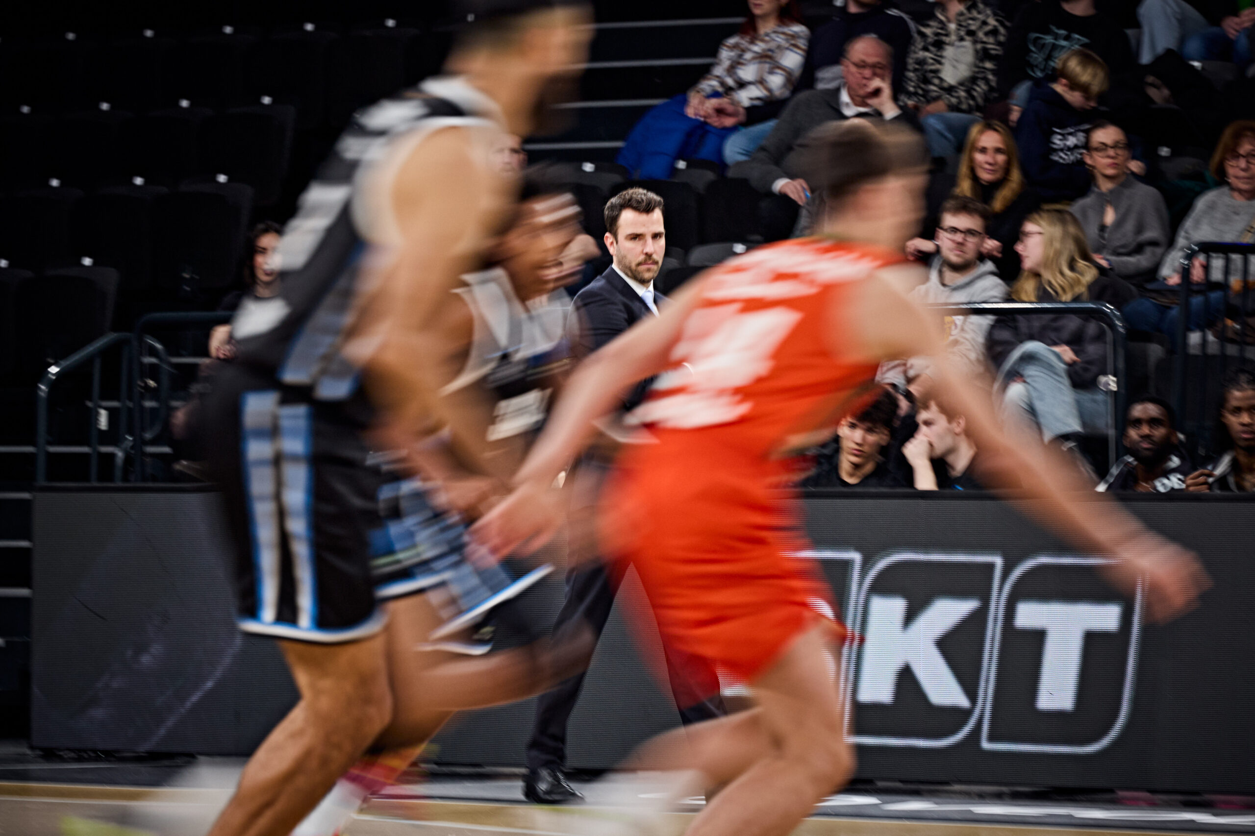 Zwei Basketballspieler in Bewegung auf dem Spielfeld, einer in schwarzem Trikot, der andere in rotem Trikot, im Hintergrund Zuschauer auf Tribünenplätzen.
