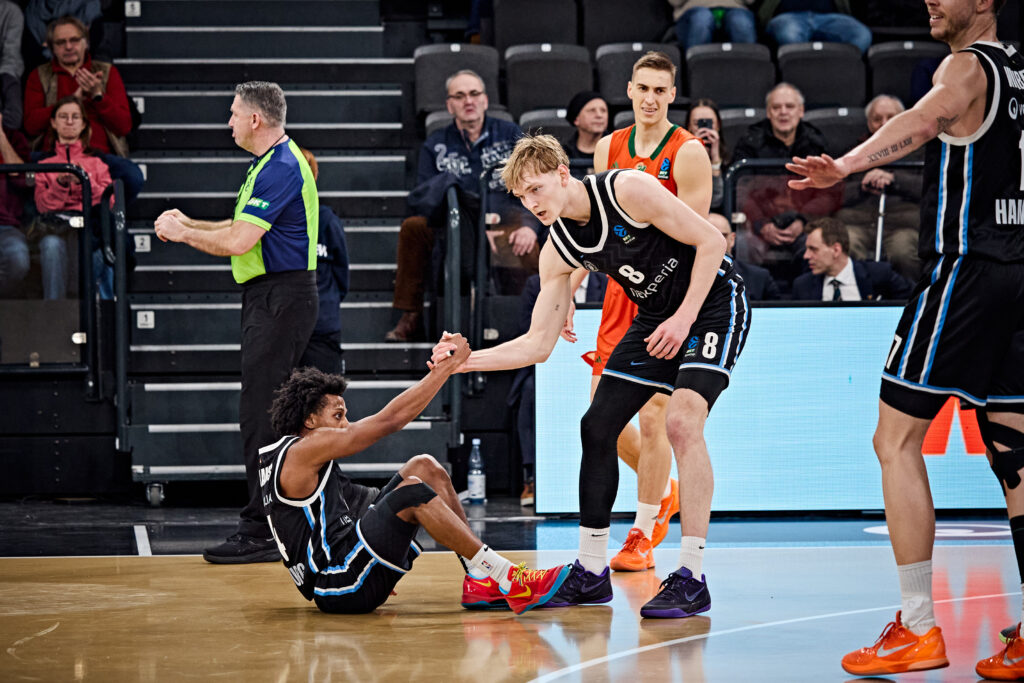 Basketballspieler in schwarzen Trikots auf dem Spielfeld, einer sitzt am Boden und wird von einem Mitspieler auf die Beine geholfen, im Hintergrund Schiedsrichter und Zuschauer.