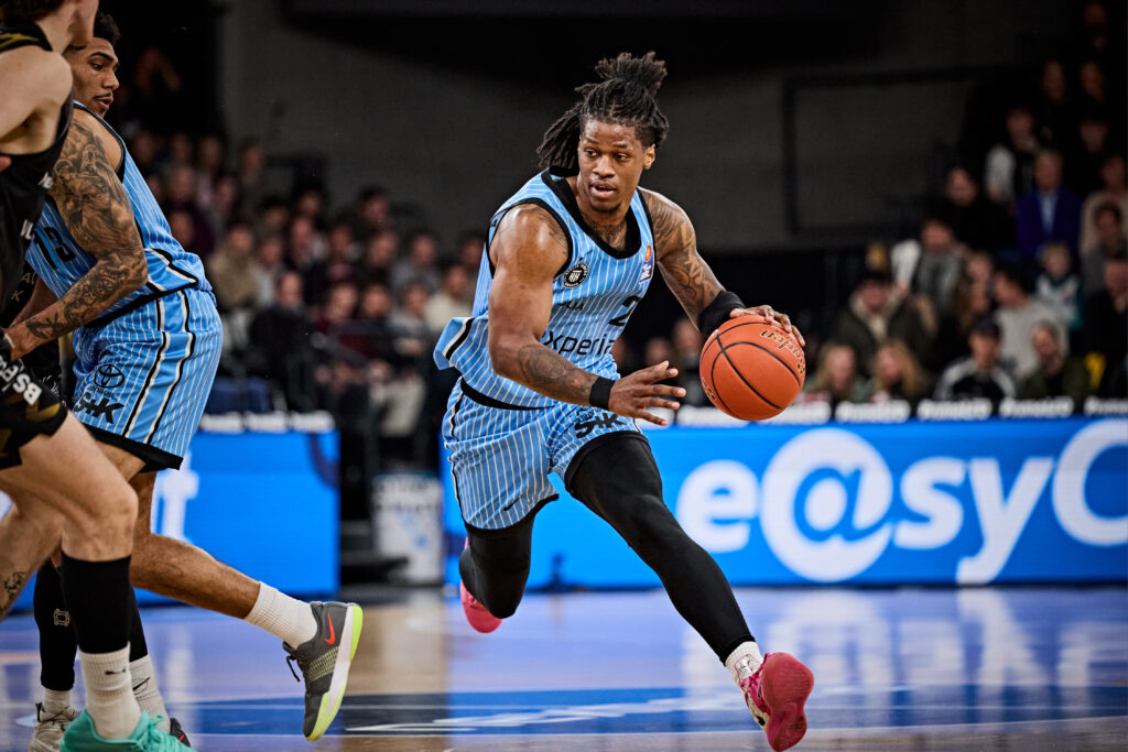 Basketballspieler in blau-schwarzem Trikot dribbelt den Ball auf dem Spielfeld vor einem Publikum