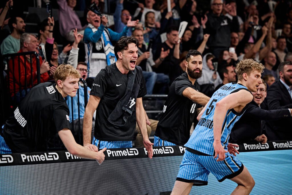 Basketballspieler in hellblauem Trikot mit der Nummer 5 läuft an der Seitenlinie an jubelnden Teammitgliedern in schwarzer Kleidung vorbei