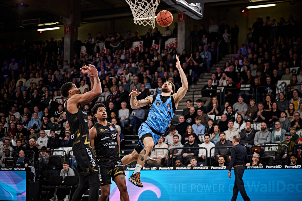 Basketballspieler in blauer Trikot springen zum Korb, umgeben von Gegnern in schwarzen Trikots, Zuschauer im Hintergrund