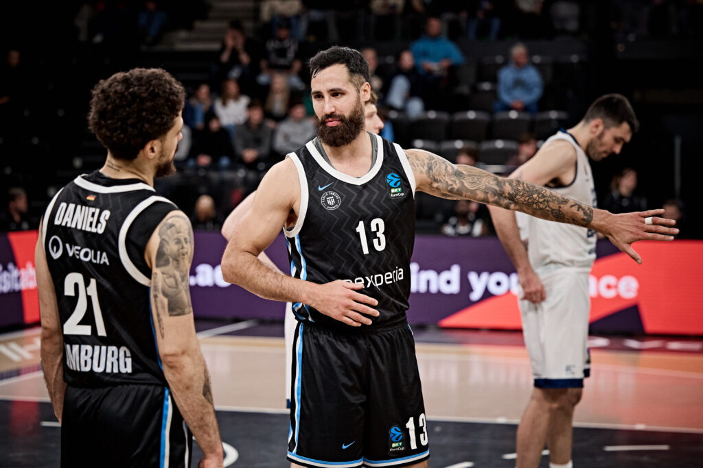 Drei Basketballspieler auf dem Spielfeld, zwei in schwarzen Trikots mit den Nummern 21 und 13, einer in weißem Trikot, einer der Spieler mit ausgestrecktem Arm und sichtbaren Tattoos am rechten Arm