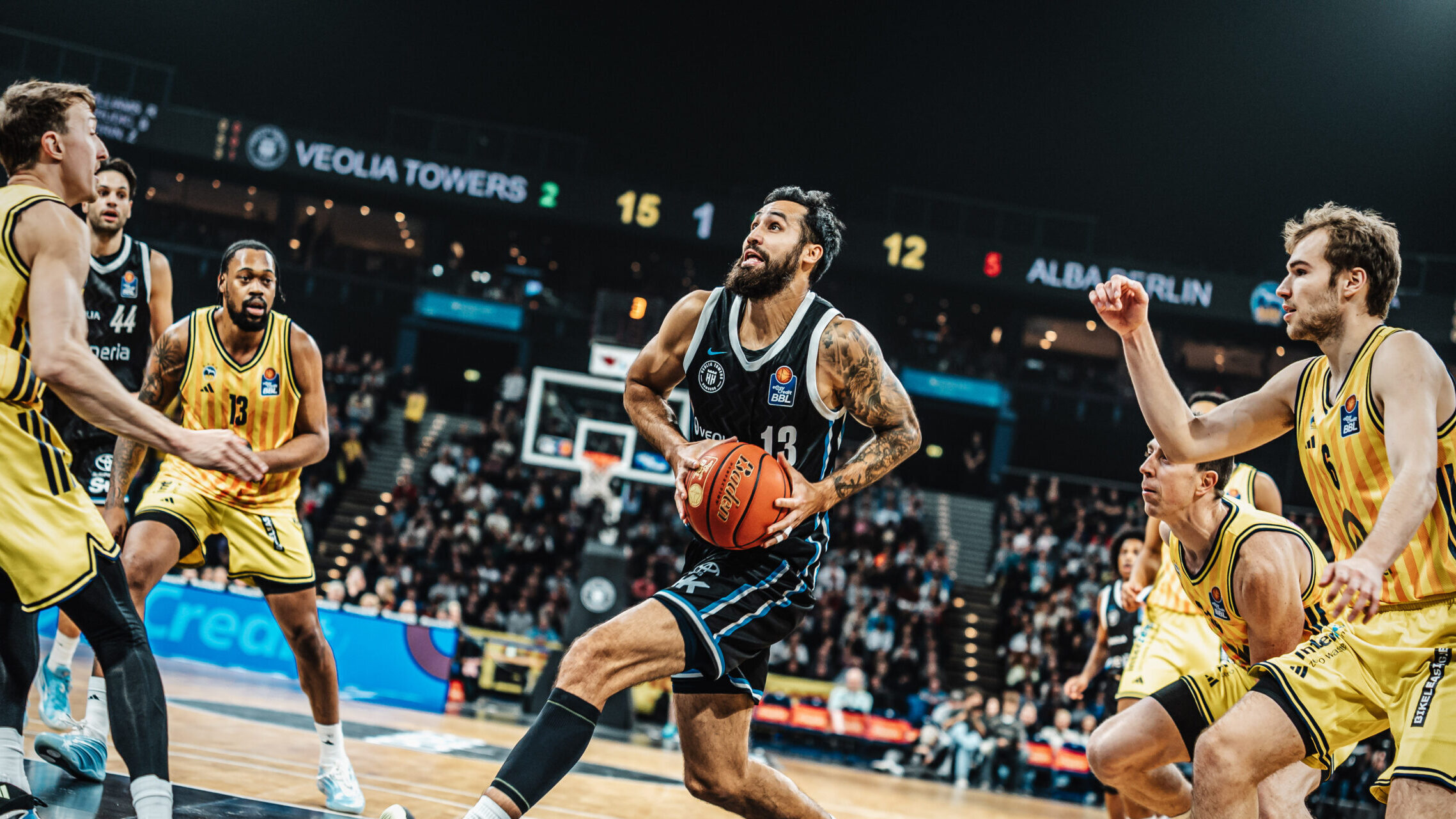 Basketballspieler in schwarzer Spielkleidung dribbelt den Ball auf dem Spielfeld, umgeben von mehreren Gegenspielern in gelber Spielkleidung