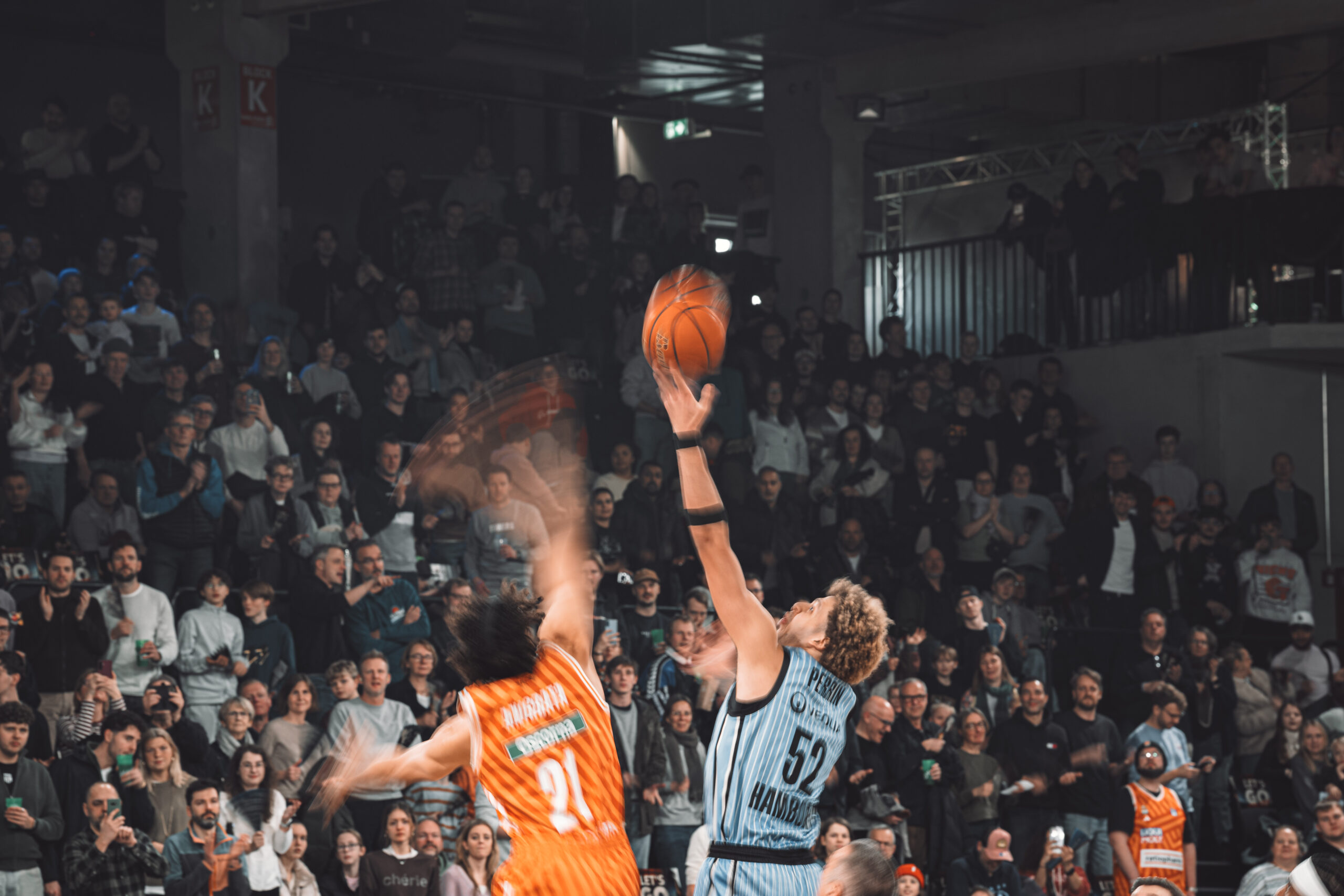 Zwei Basketballspieler springen zum Ball, einer in orangefarbenem Trikot mit der Nummer 21, der andere in hellblauem Trikot mit der Nummer 52, vor einer Zuschauertribüne in einer Halle