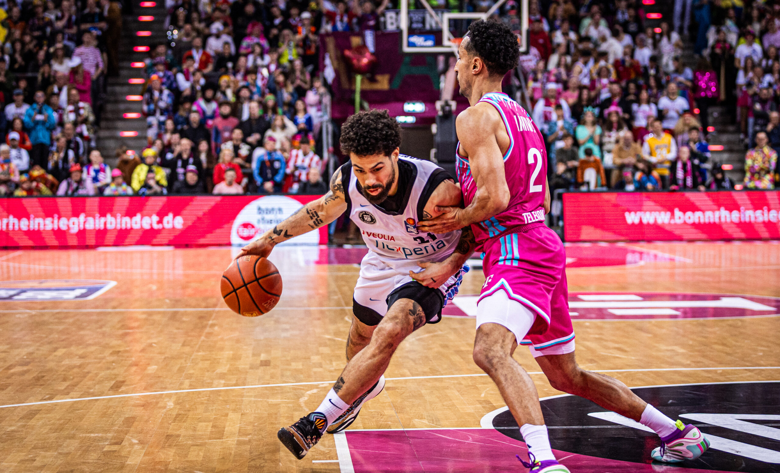 Zwei Basketballspieler auf dem Spielfeld einer Halle, einer dribbelt den Ball, der andere verteidigt mit ausgestrecktem Arm