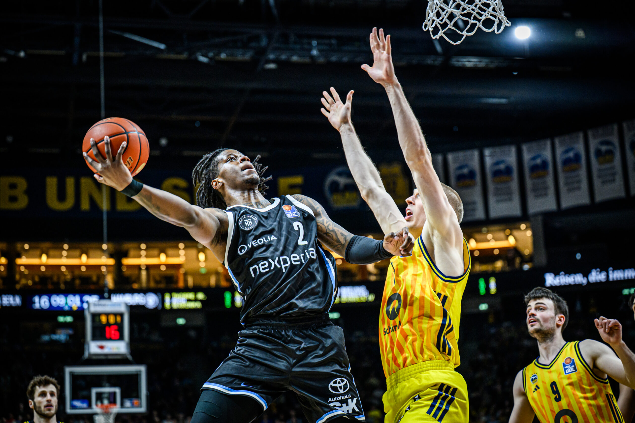 Basketballspieler in schwarzem Trikot mit Ball in der linken Hand springt zum Korb, während zwei Spieler in gelben Trikots ihn verteidigen