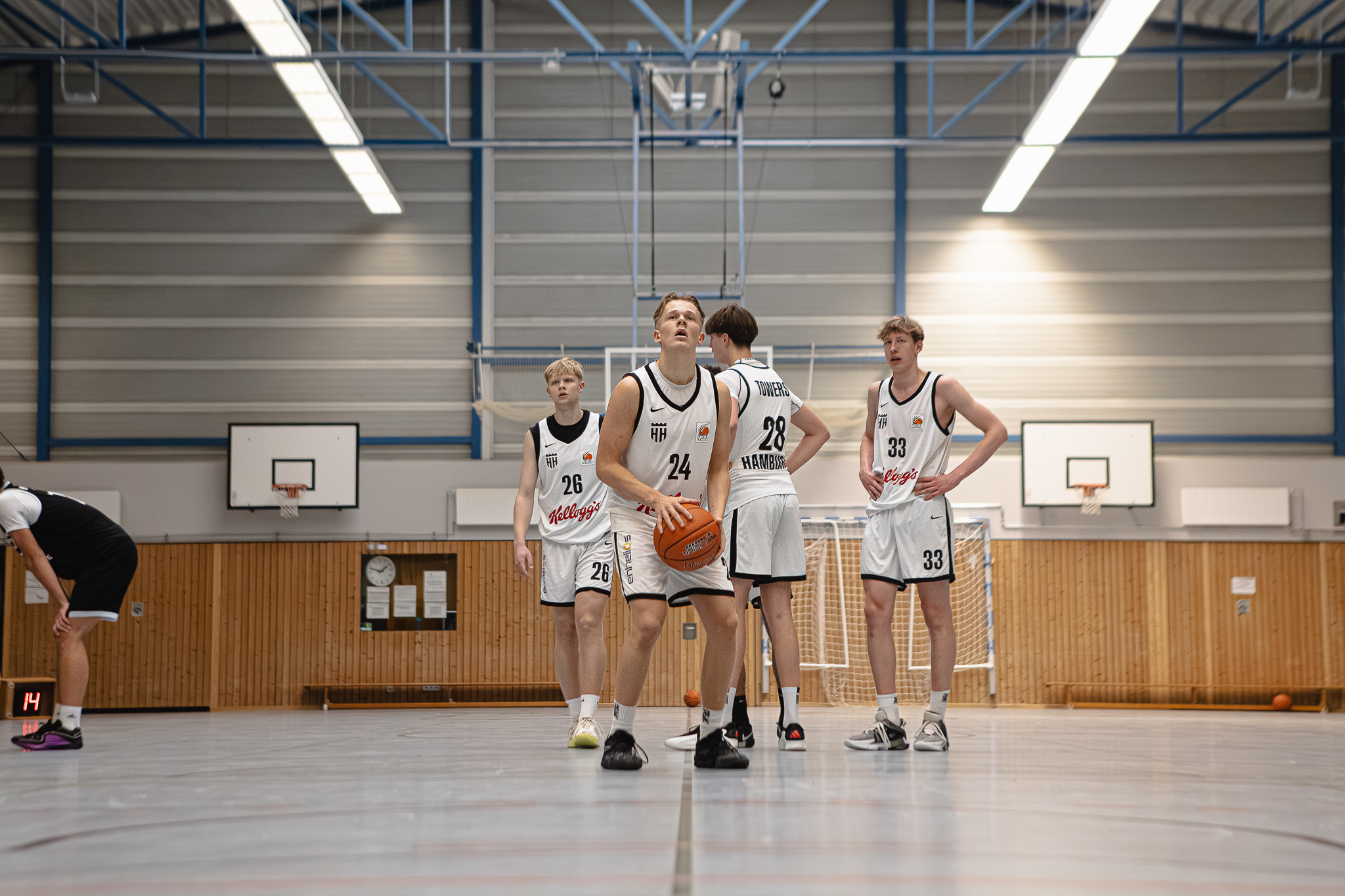 Vier Basketballspieler in weißen Trikots mit schwarzen Nummern stehen auf einem Hallenboden vor zwei Basketballkörben