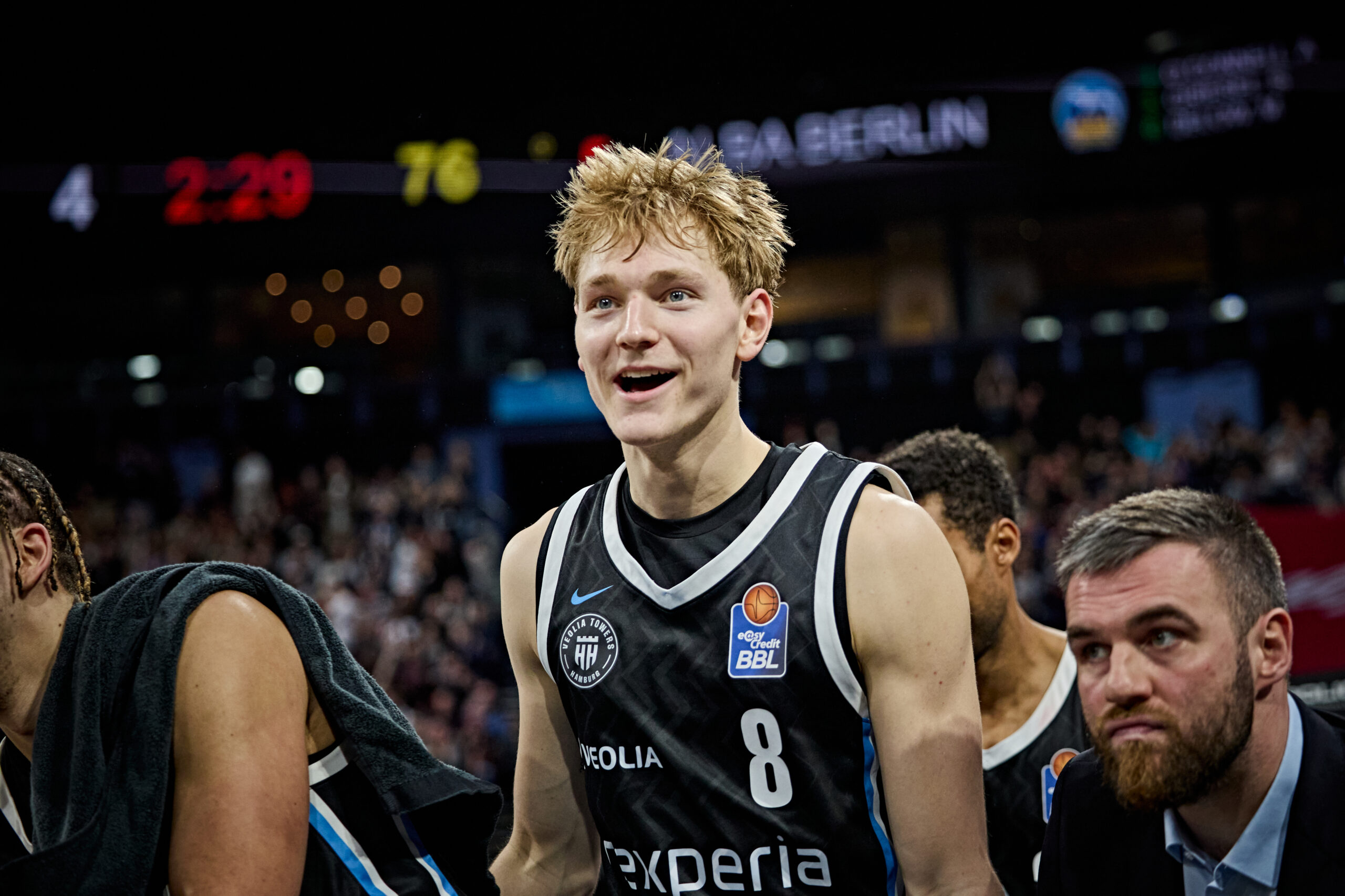 Mehrere Basketballspieler in schwarzen Trikots mit der Aufschrift "nexperia" und der Nummer 8 im Vordergrund in einer Halle mit Publikum im Hintergrund