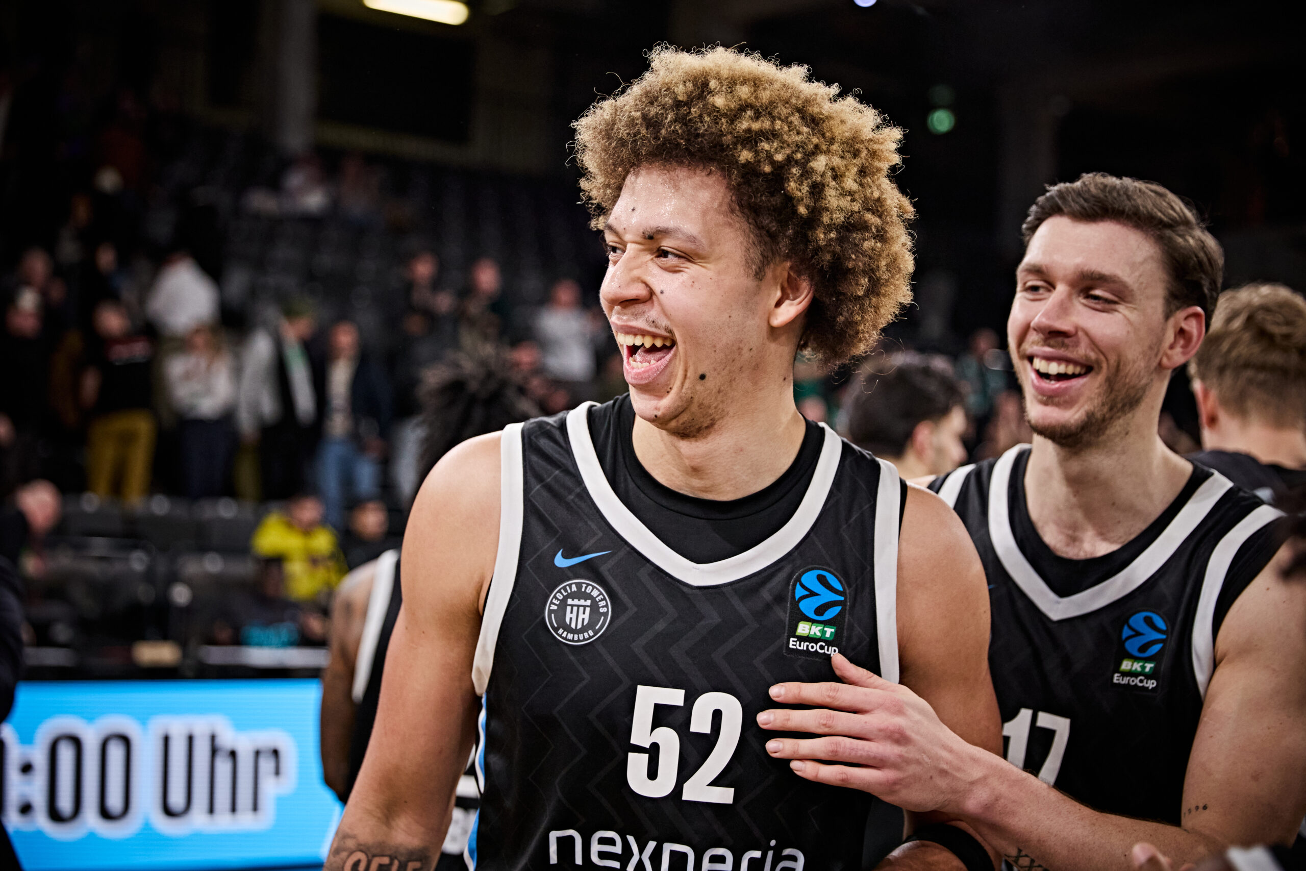 Zwei Basketballspieler in schwarzen Trikots mit weißen Rändern und EuroCup-Logo, einer trägt die Nummer 52, der andere die Nummer 17, im Hintergrund Zuschauer in einer Halle