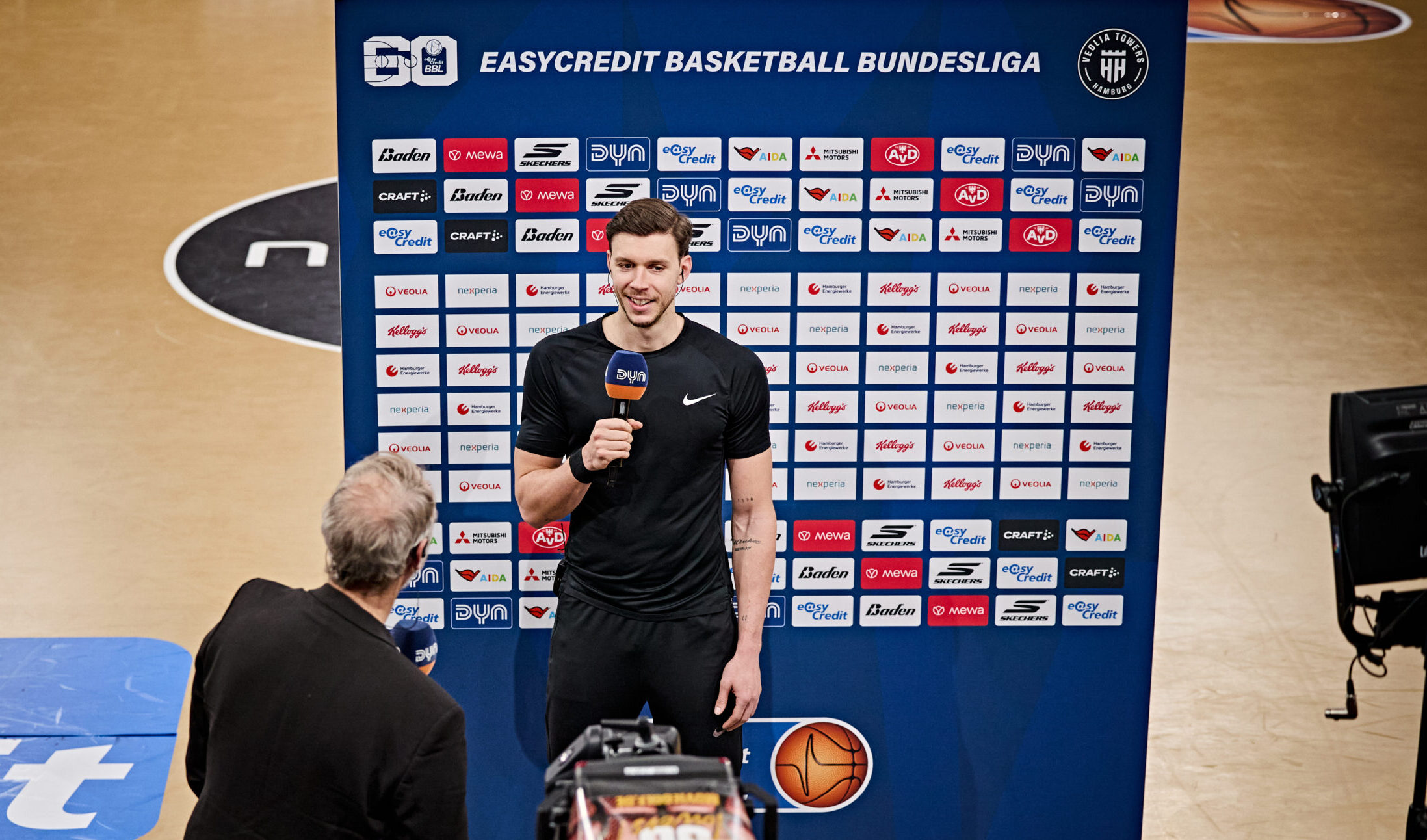 Ein Mann in schwarzem Sportoutfit steht vor einer Sponsorenwand und hält ein Mikrofon, vor ihm steht ein Interviewer mit Mikrofon und eine Kamera ist auf sie gerichtet.