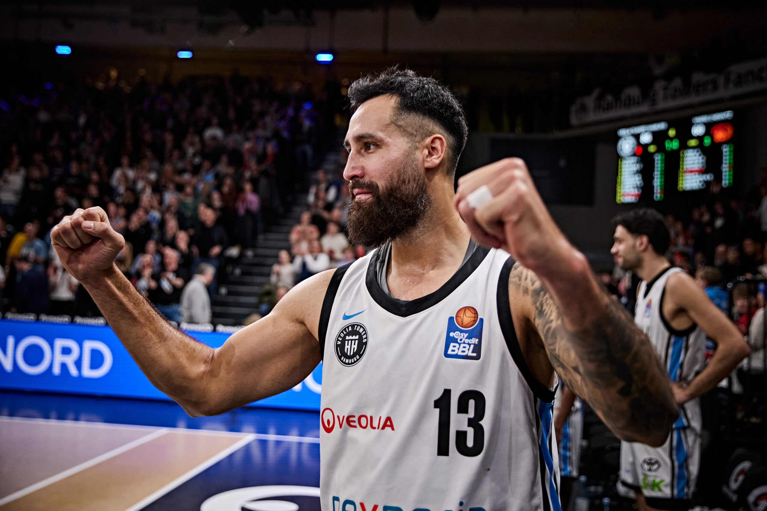 Basketballspieler mit Trikotnummer 13 hebt die Fäuste in einer Halle mit Publikum