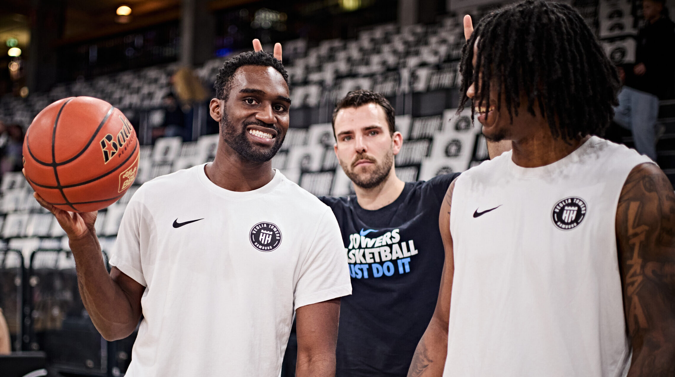 Drei Basketballspieler in Sportkleidung stehen in einer Halle, einer hält einen Basketball in der Hand