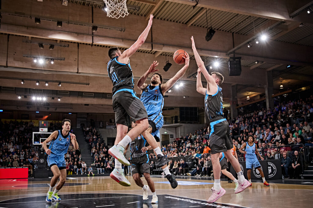 Mehrere Basketballspieler in blauen und schwarzen Trikots springen in einer Halle mit Publikum und versuchen, den Ball zu erobern