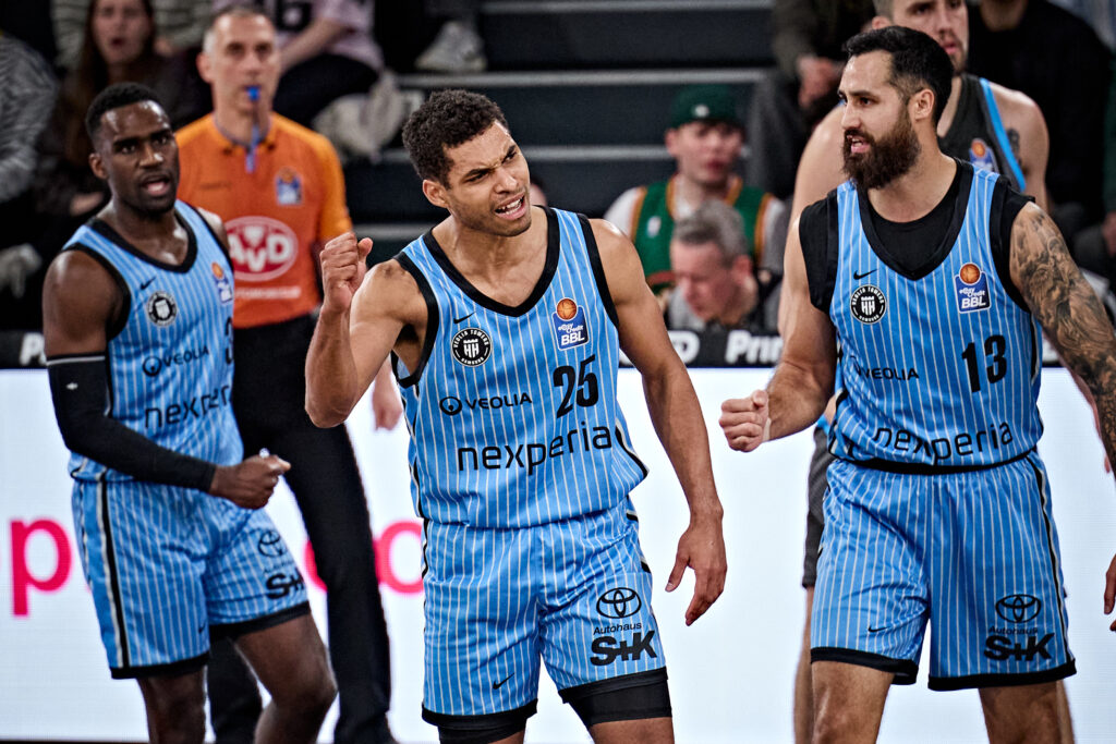 Drei Basketballspieler in blau-schwarzen Trikots mit senkrechten Streifen auf dem Spielfeld in Jubelpose