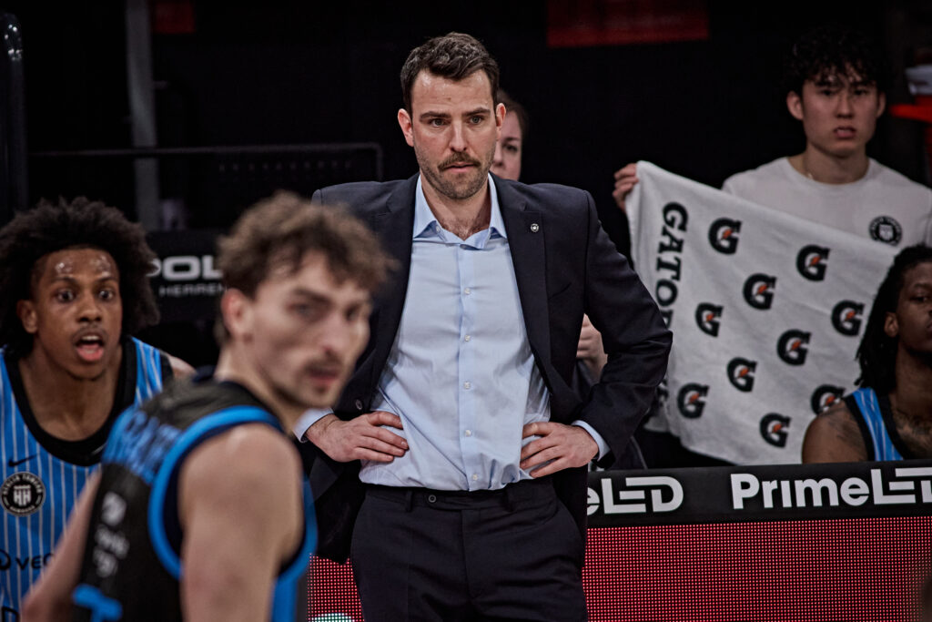 Mann in Anzug mit verschränkten Händen vor dem Bauch steht vor Basketballspielern in Trikots, im Hintergrund Person mit Tuch mit Gatorade-Logo
