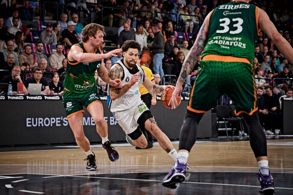 Drei Basketballspieler in Aktion auf dem Spielfeld, zwei in grünen Trikots mit der Aufschrift 'VET-CONCEPT GLADIATORS TRIER', einer in weißem Trikot mit Ball, Zuschauer im Hintergrund