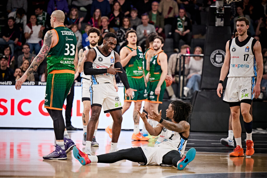Mehrere Basketballspieler in weißen und grünen Trikots auf dem Spielfeld, ein Spieler in weiß sitzt auf dem Boden und gestikuliert