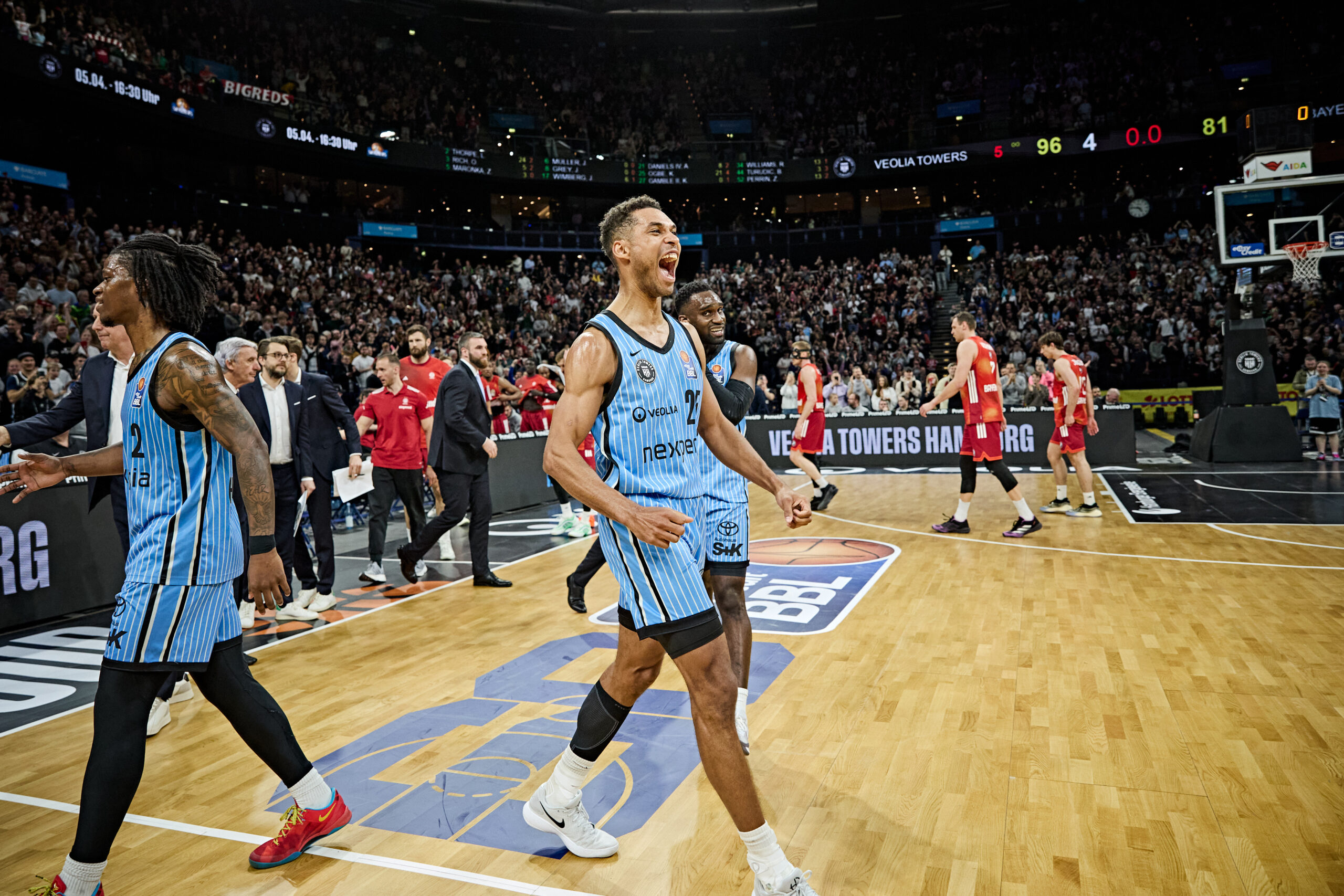 Basketballspieler in hellblauen Trikots verlassen das Spielfeld vor einer jubelnden Zuschauermenge in einer Halle