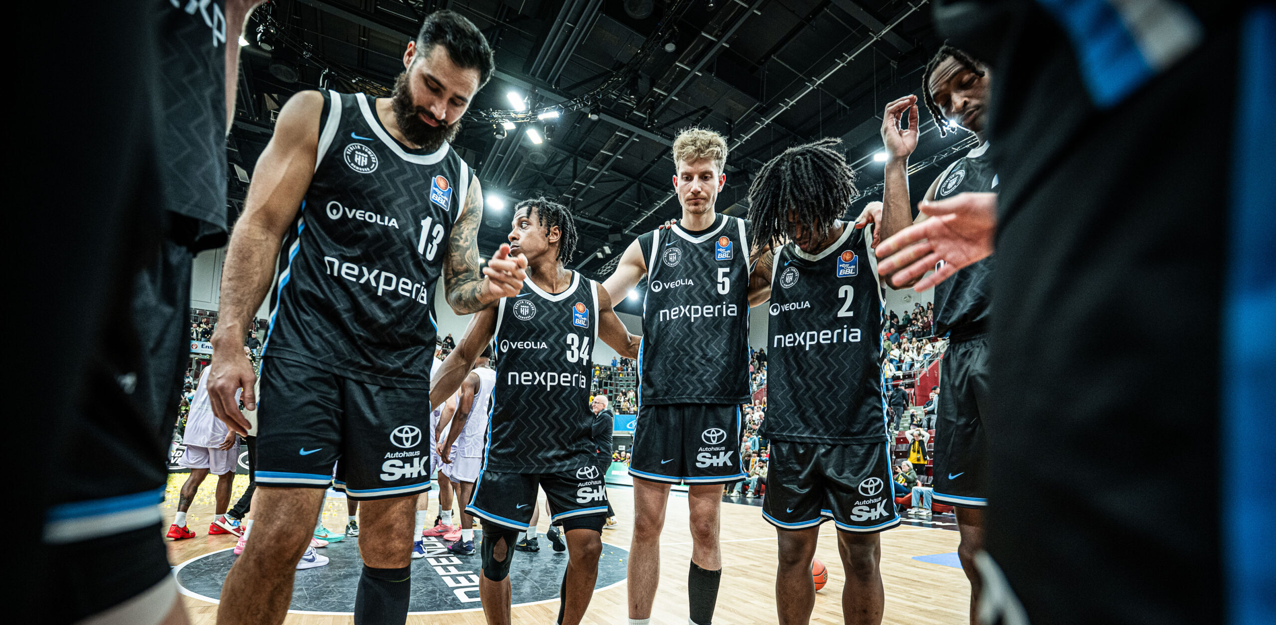 Fünf Basketballspieler in schwarzen Trikots mit den Aufschriften "nexperia" und "Toyota" stehen auf dem Spielfeld und halten sich an den Händen.