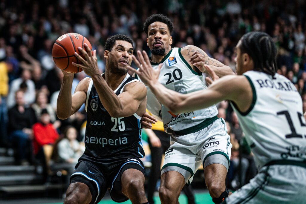 Drei Basketballspieler in Aktion, zwei in weißen Trikots mit den Nummern 20 und 14 verteidigen gegen einen Spieler in schwarzem Trikot mit der Nummer 25, der den Ball hält