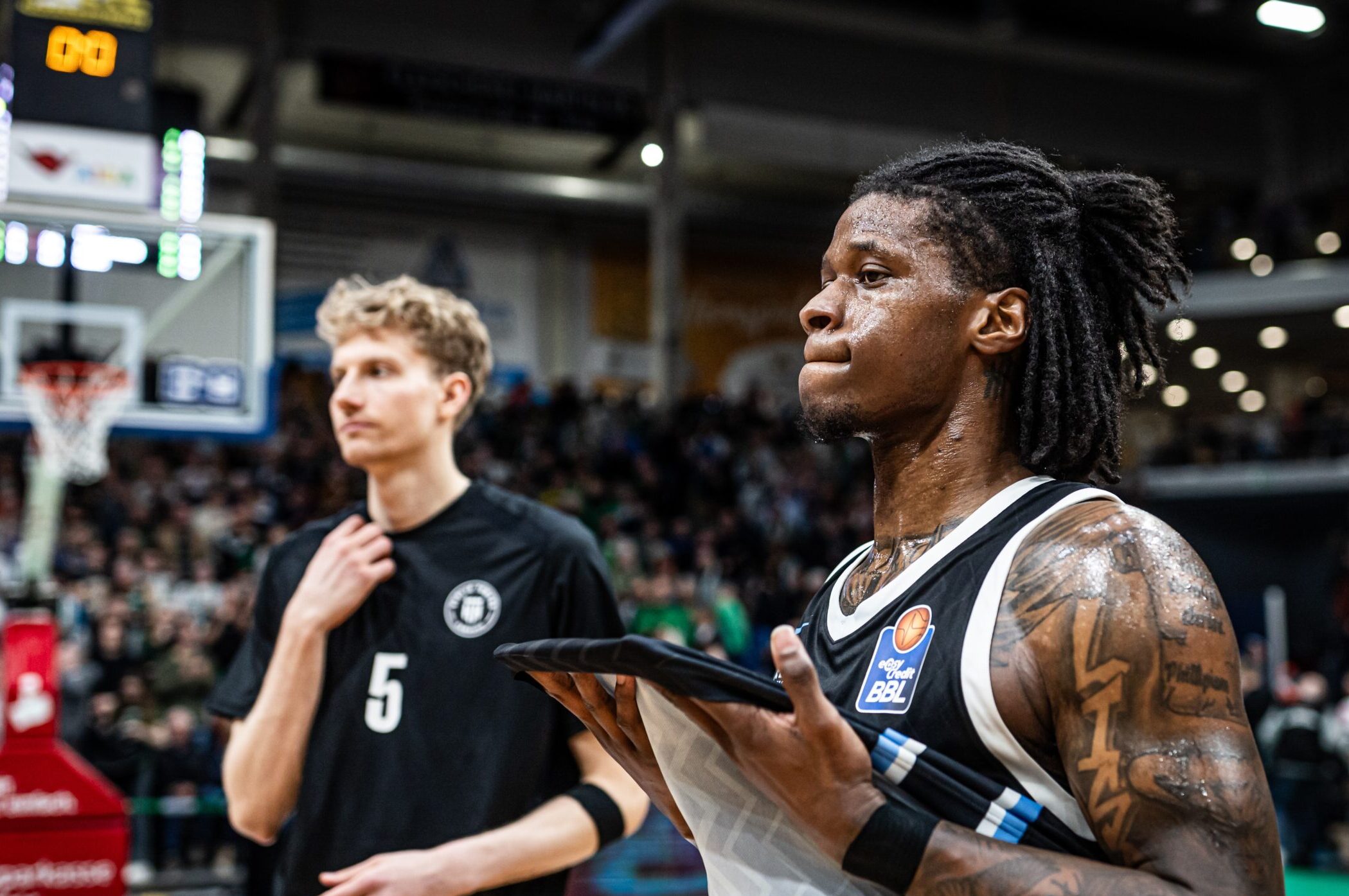 Zwei Basketballspieler auf dem Spielfeld, einer mit Tattoos und Trikot mit BBL-Logo, der andere mit schwarzem Trikot und der Nummer 5