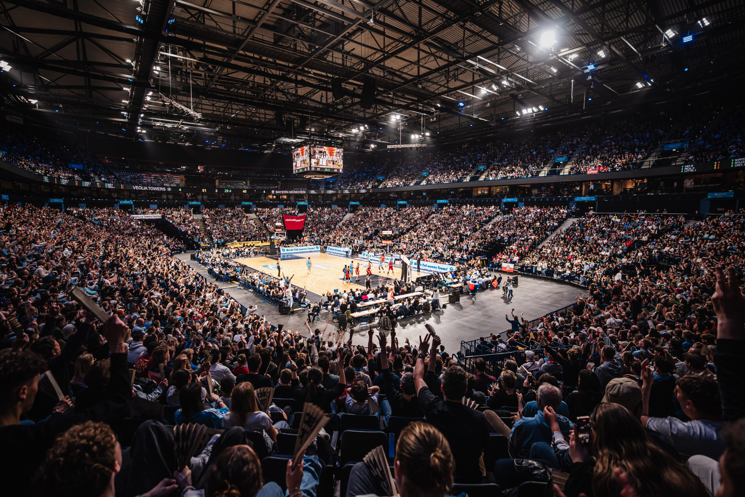 Voll besetzte Basketballarena mit Spielern auf dem Spielfeld und jubelnden Zuschauern