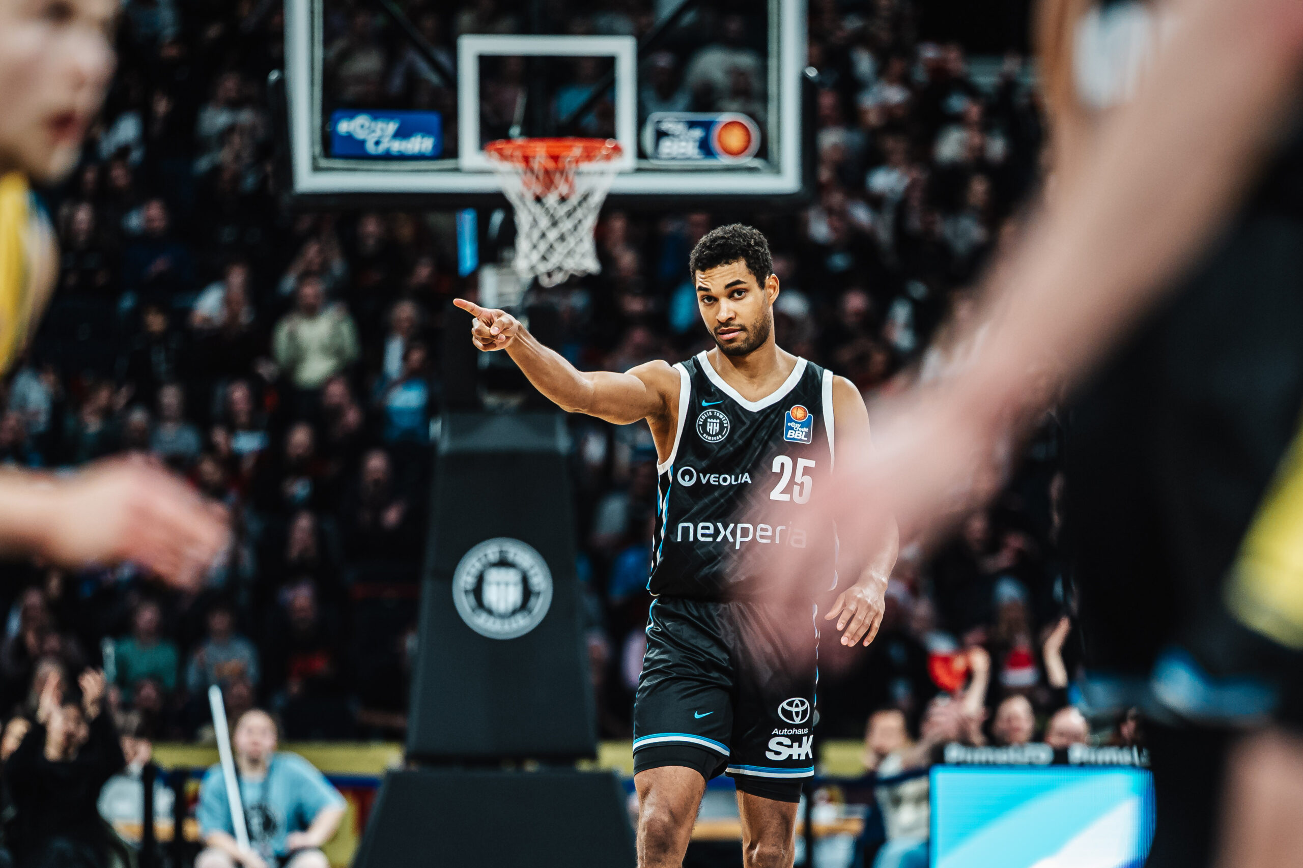 Basketballspieler mit Trikotnummer 25 zeigt mit ausgestrecktem Arm auf dem Spielfeld vor einem Basketballkorb