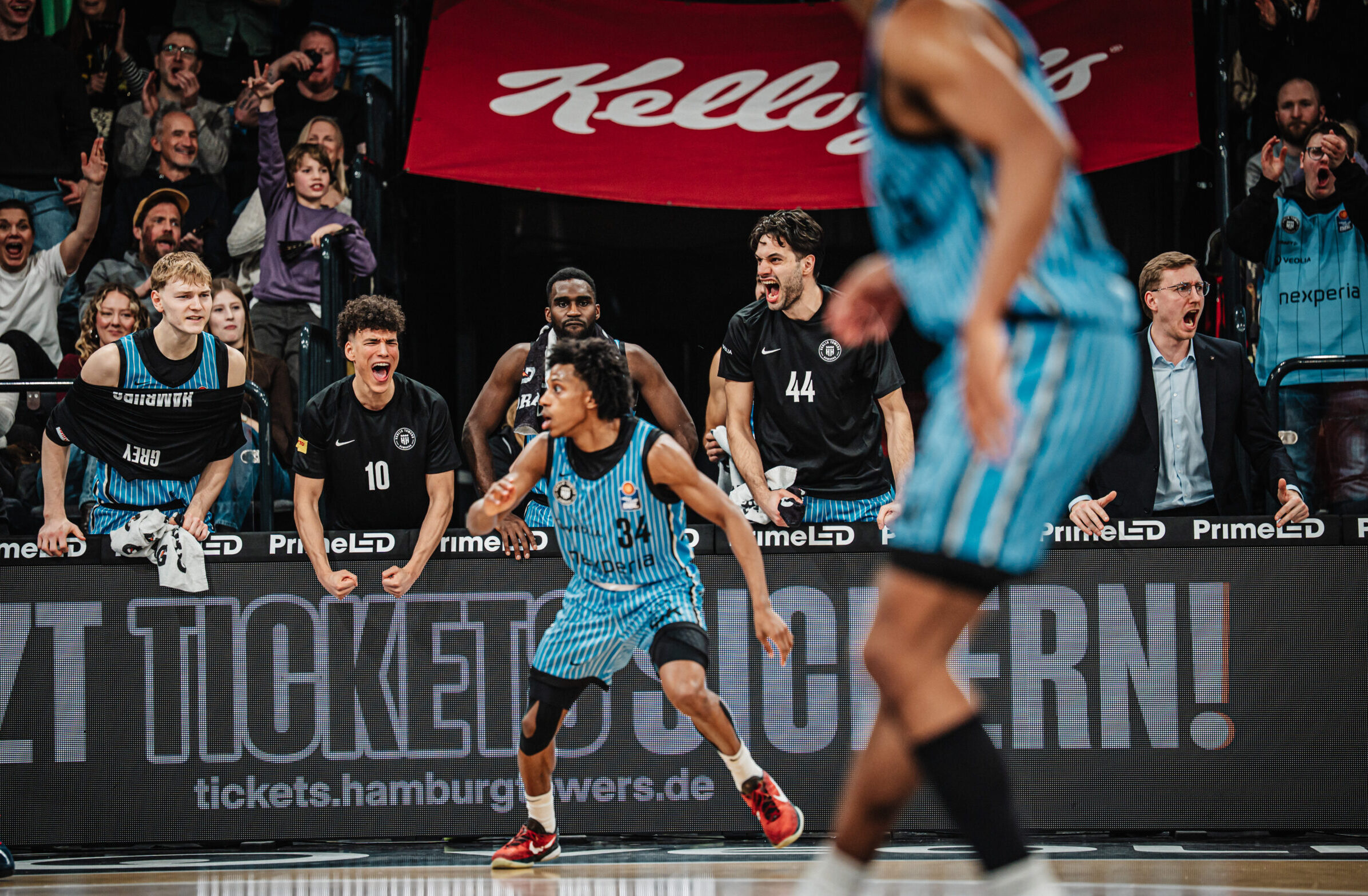 Mehrere Basketballspieler in Aktion auf dem Spielfeld vor einer Zuschauertribüne mit Werbebannern.