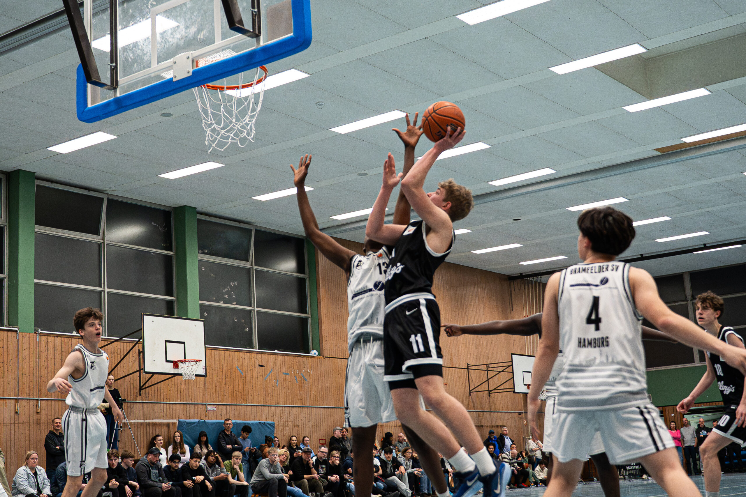 Mehrere Basketballspieler in Aktion unter dem Korb in einer Halle mit Zuschauern auf der Tribüne