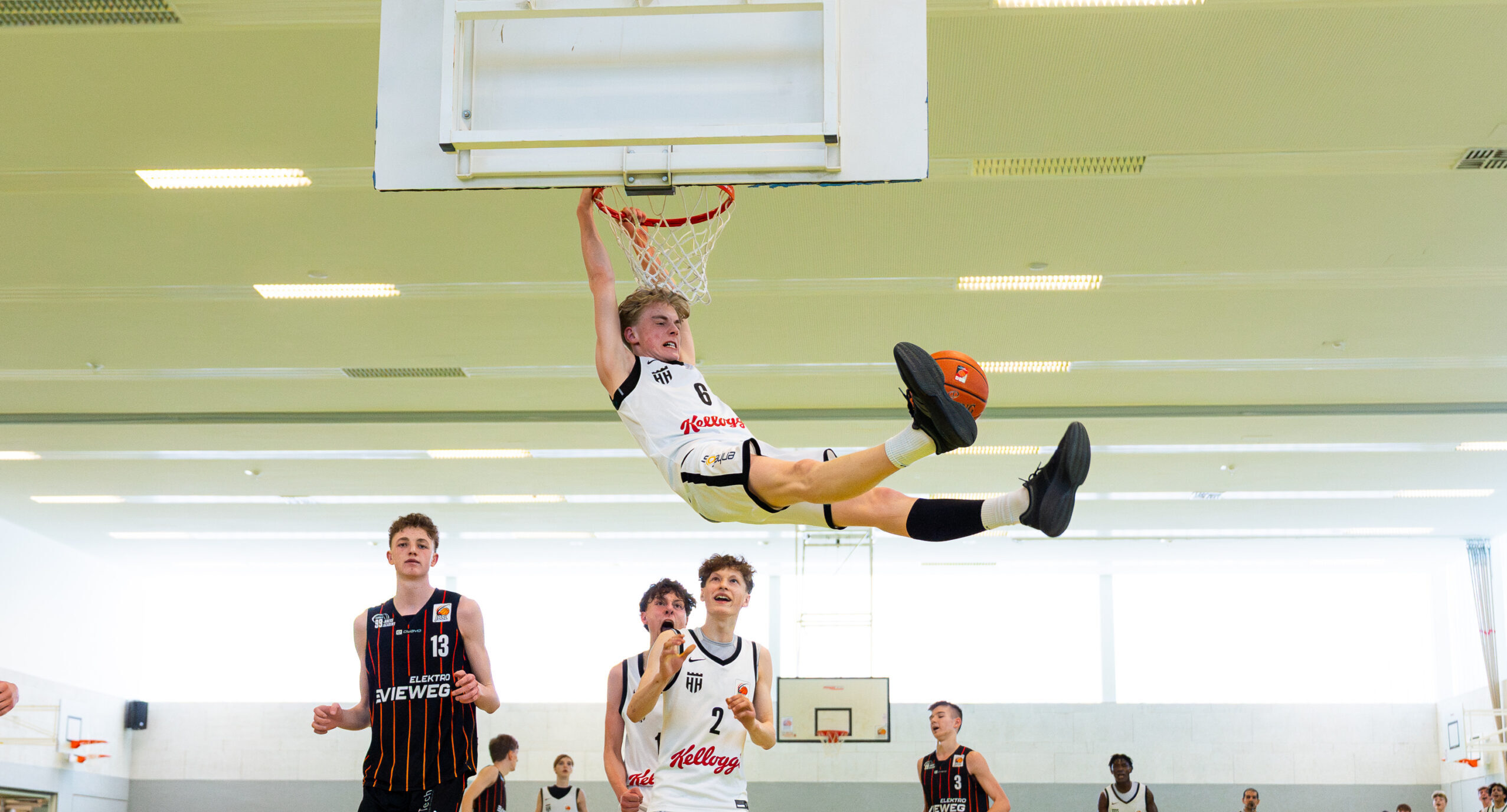 Ein Basketballspieler hängt mit ausgestreckten Beinen am Korb nach einem Dunking, weitere Spieler beobachten das Geschehen in der Halle