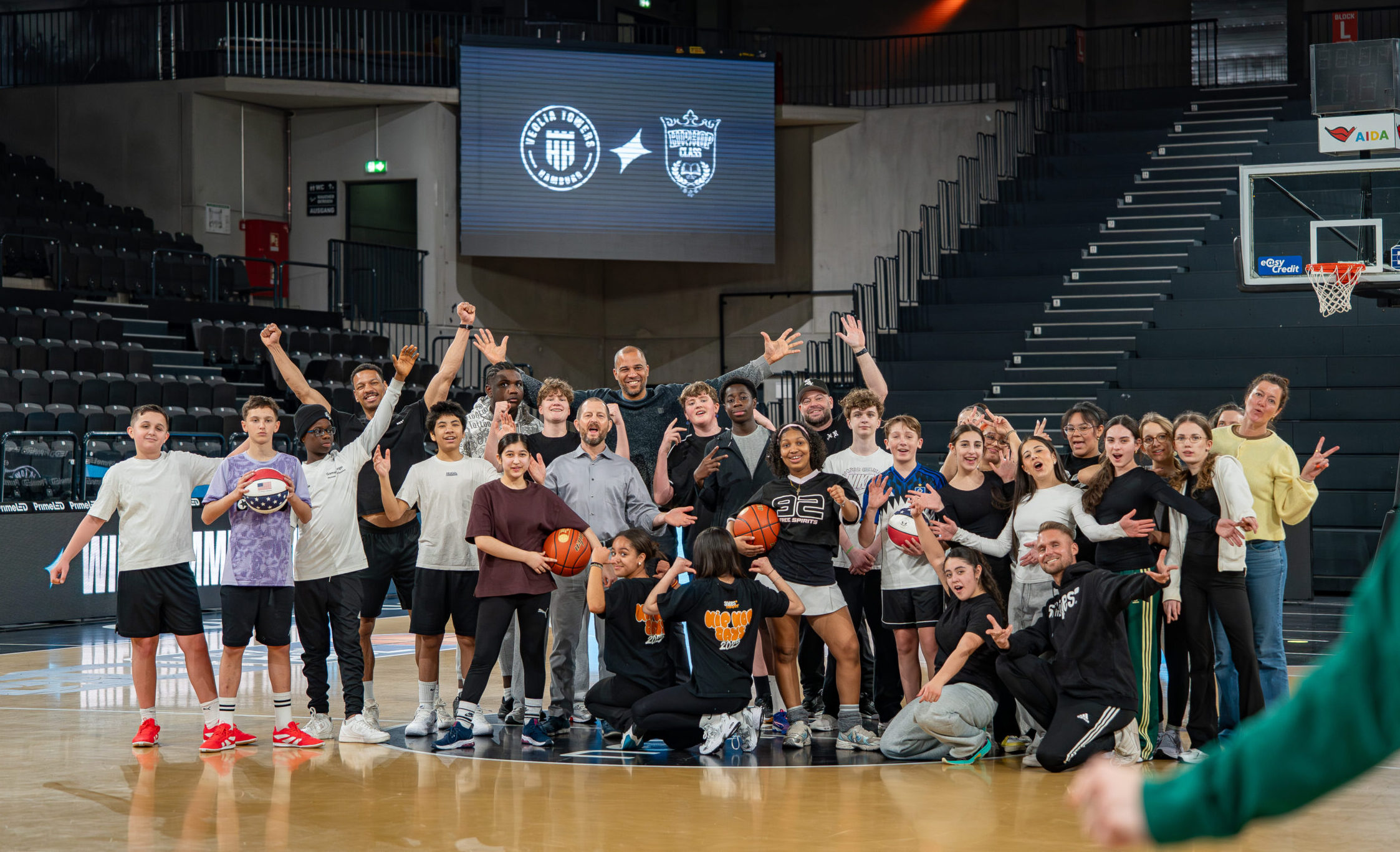 Gruppe von Jugendlichen und Erwachsenen auf einem Basketballfeld in einer Halle posiert mit Basketbällen und hebt die Arme