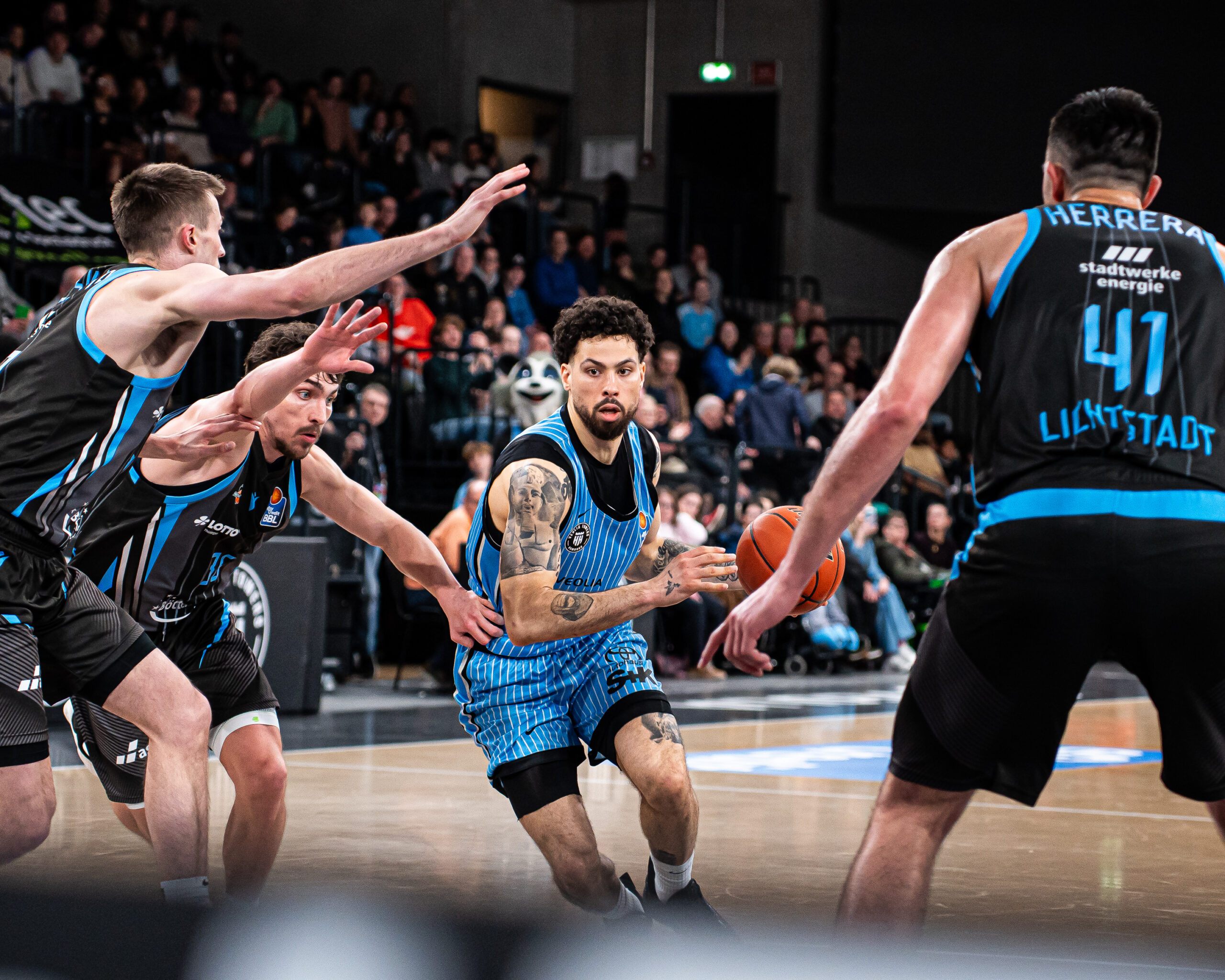 Basketballspieler in blauen und schwarzen Trikots kämpfen um den Ball auf dem Spielfeld vor Zuschauern