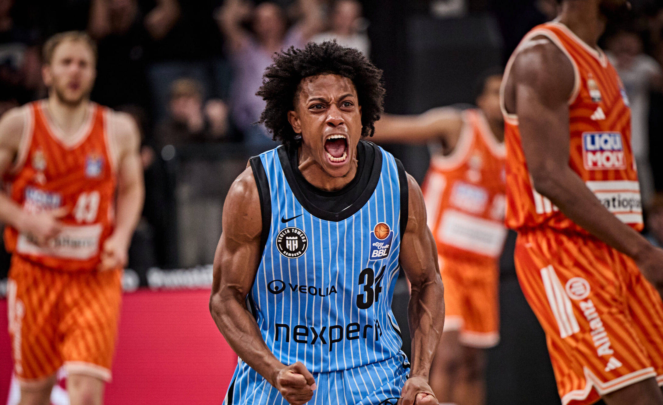Basketballspieler in blau-weiß gestreiftem Trikot mit geballten Fäusten auf dem Spielfeld, umgeben von Spielern in orangefarbenen Trikots