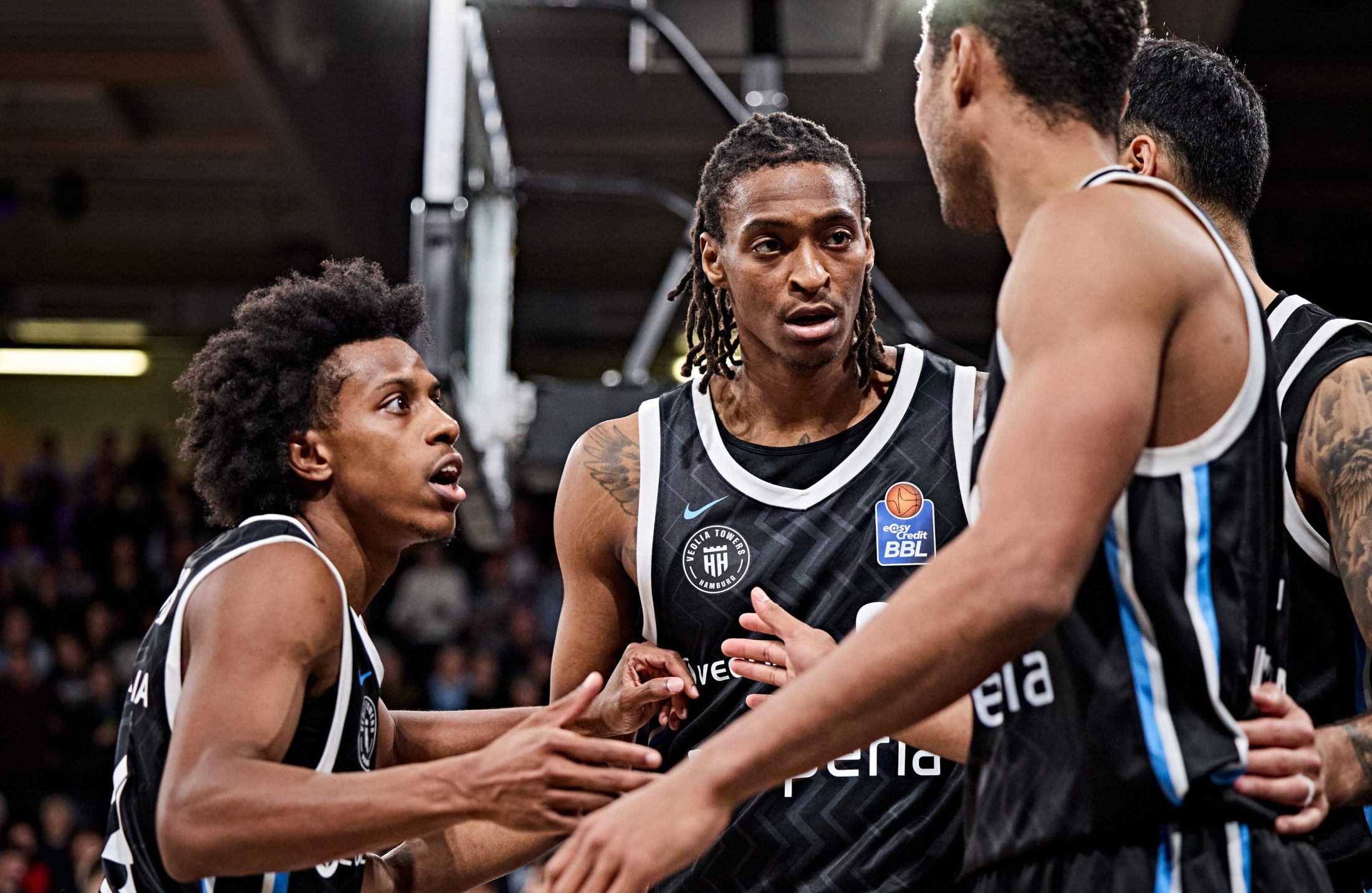 Vier Basketballspieler in schwarzen Trikots mit weißen und blauen Streifen stehen eng beieinander und berühren sich an den Händen im Spielgeschehen