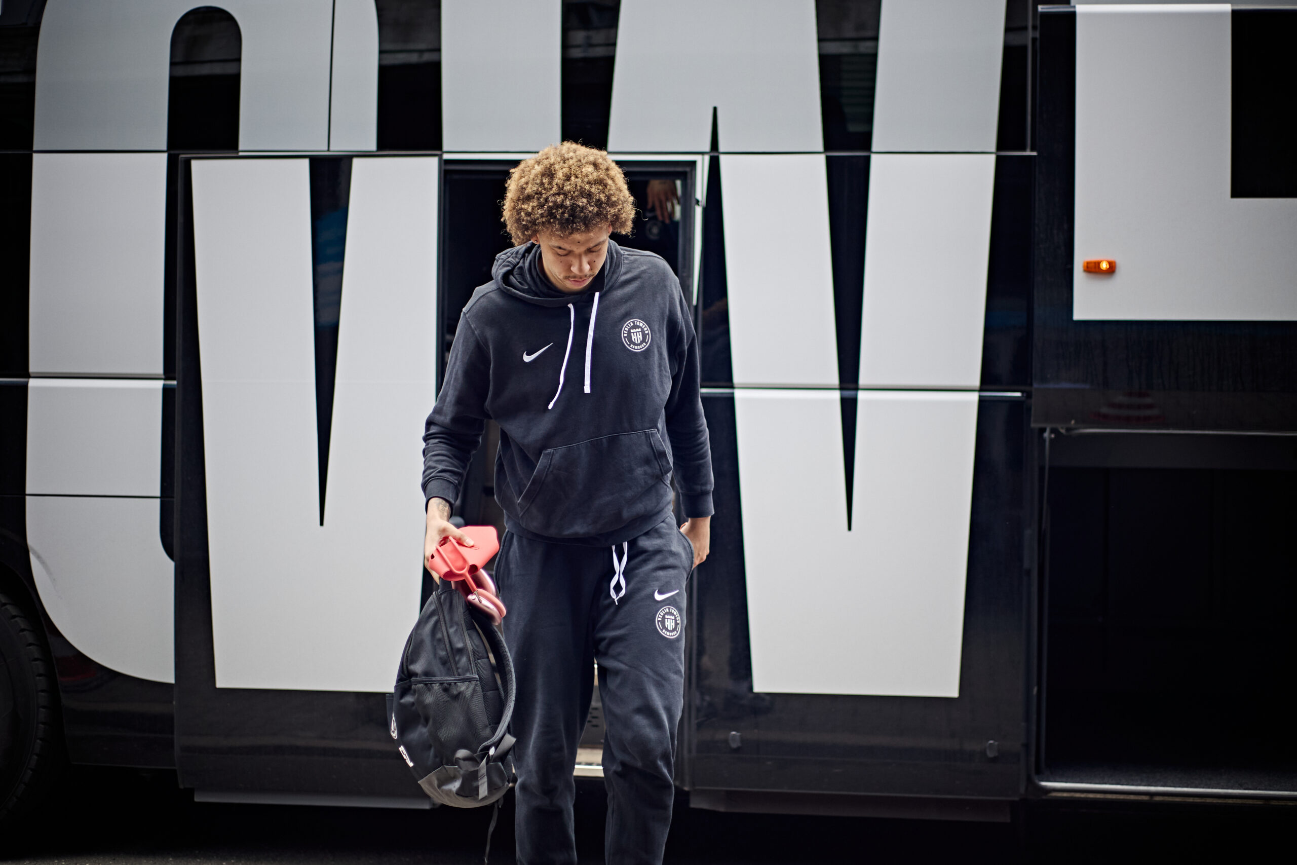 Junger Mann mit lockigem Haar in Sportbekleidung mit Vereinslogo geht vor einem Bus mit großen Buchstaben auf der Seite