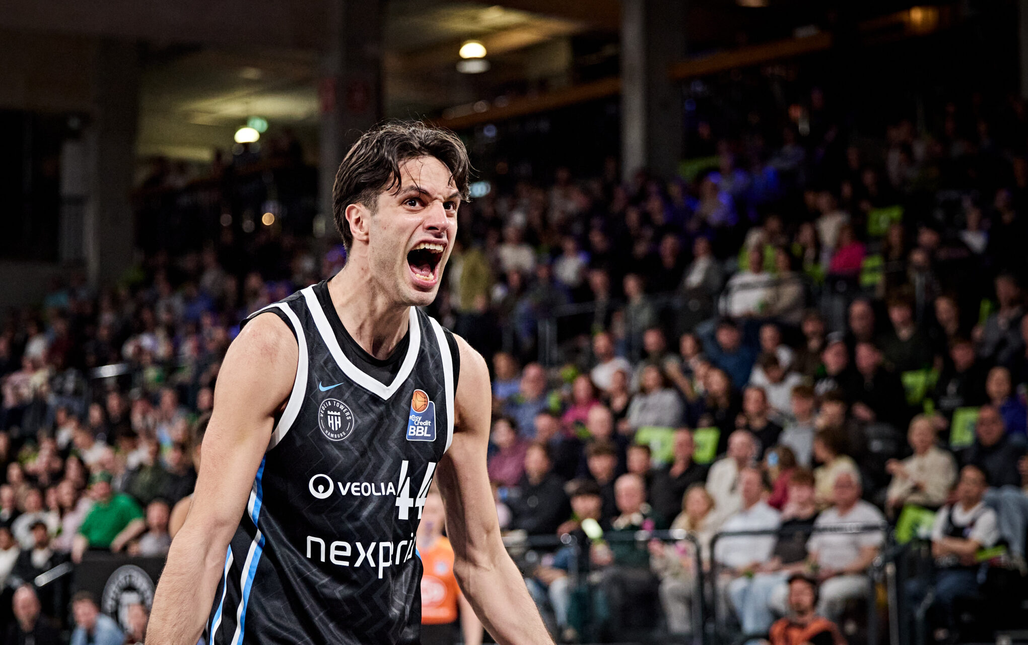 Basketballspieler in schwarzem Trikot mit weißen und blauen Akzenten steht mit angespannten Armen vor Zuschauerrängen