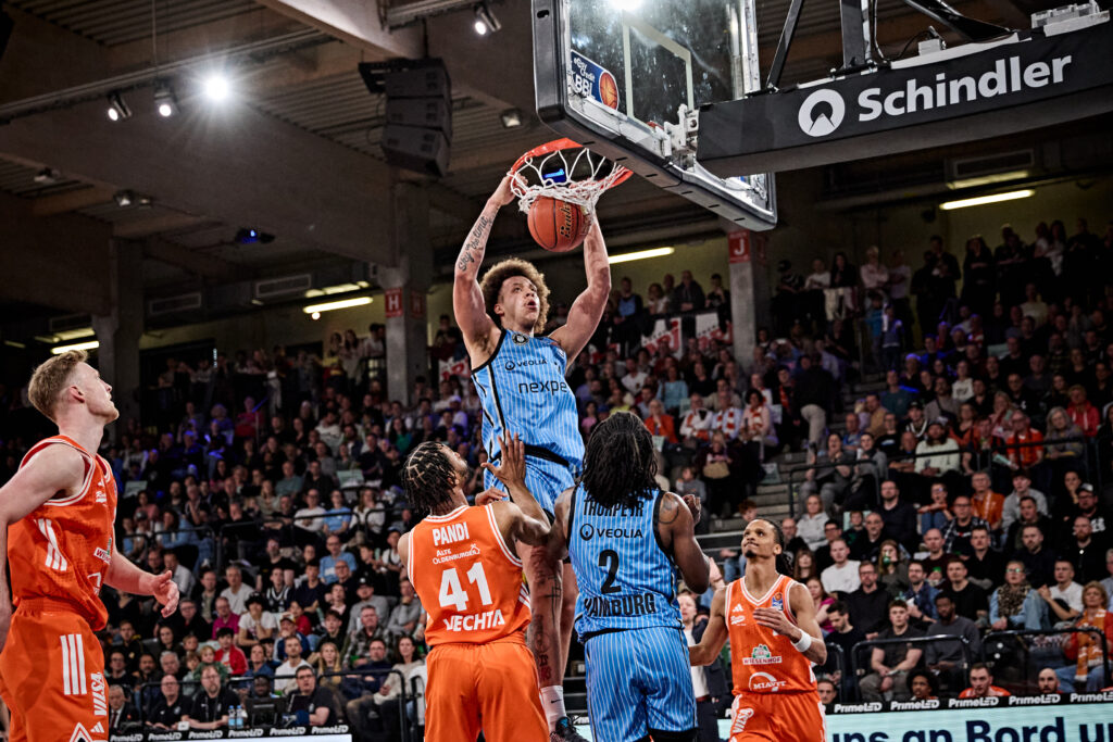 Basketballspieler in blauer Trikotnummer 7 springt zum Korb für einen Dunk, umgeben von Spielern in orangen Trikots und Zuschauern in der Halle