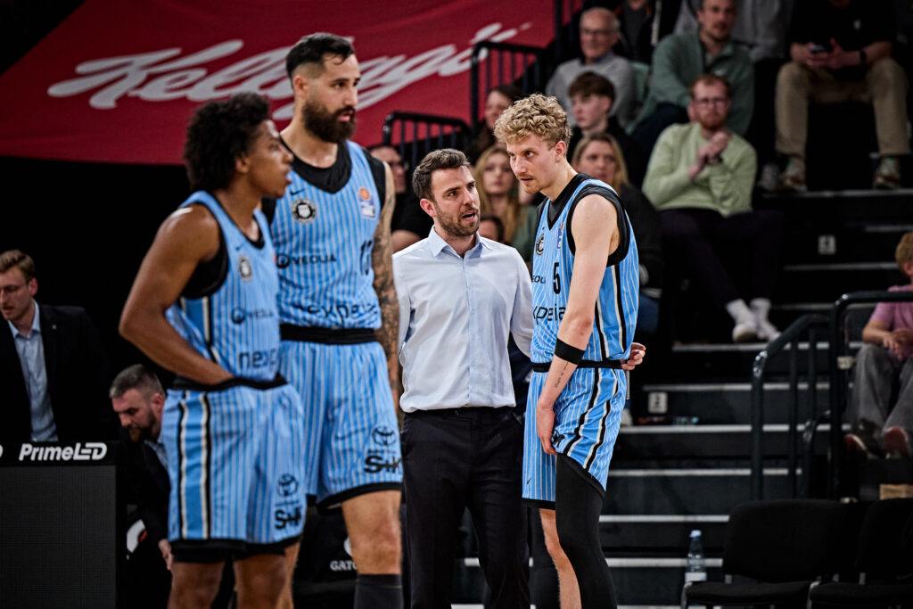 Vier Basketballspieler in hellblauen Trikots und ein Mann in weißem Hemd stehen in einer Sporthalle und unterhalten sich