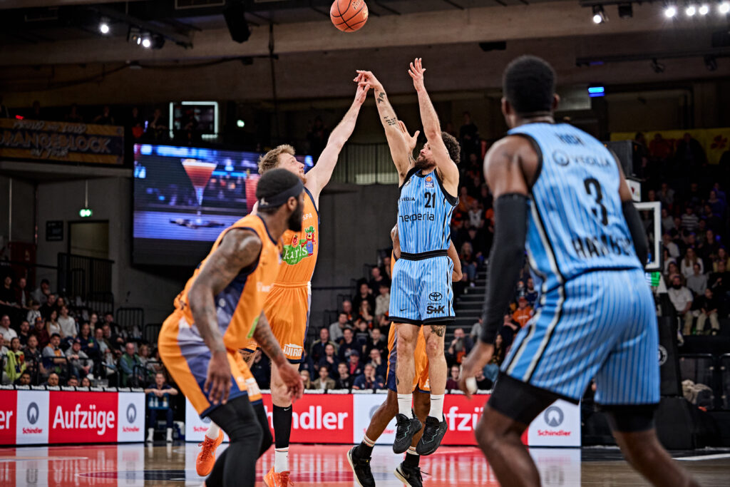 Zwei Basketballspieler springen zum Wurf, umgeben von weiteren Spielern und Zuschauern in der Halle