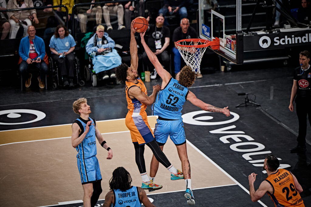 Zwei Basketballspieler springen zum Ball unter dem Korb, einer in orange, einer in blau, weitere Spieler und Zuschauer im Hintergrund
