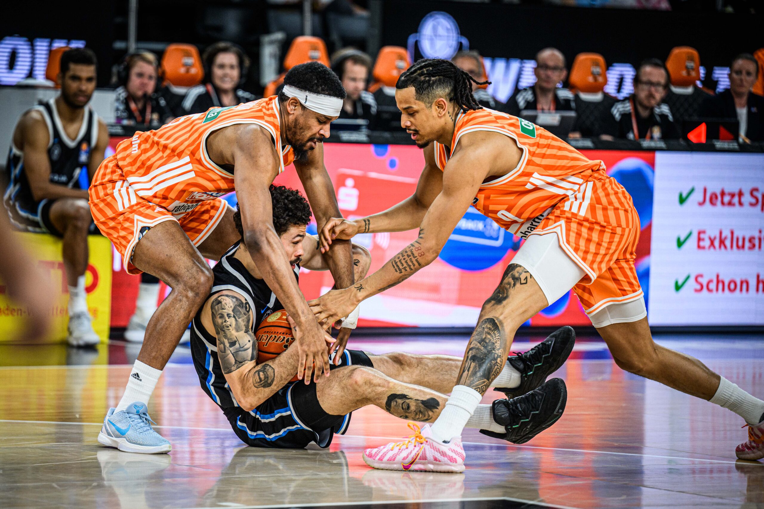 Zwei Basketballspieler in orange-weiß gestreiften Trikots greifen nach dem Ball eines am Boden liegenden Gegenspielers in schwarzem Trikot