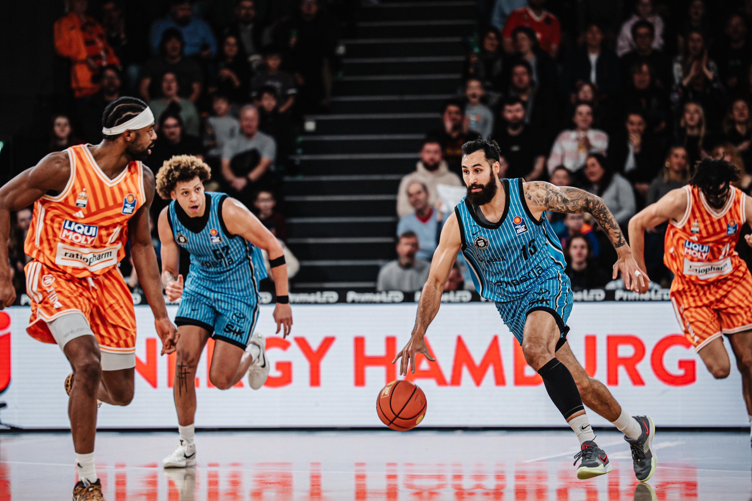 Basketballspieler in blauen und orange-weißen Trikots laufen auf dem Spielfeld, einer dribbelt den Ball